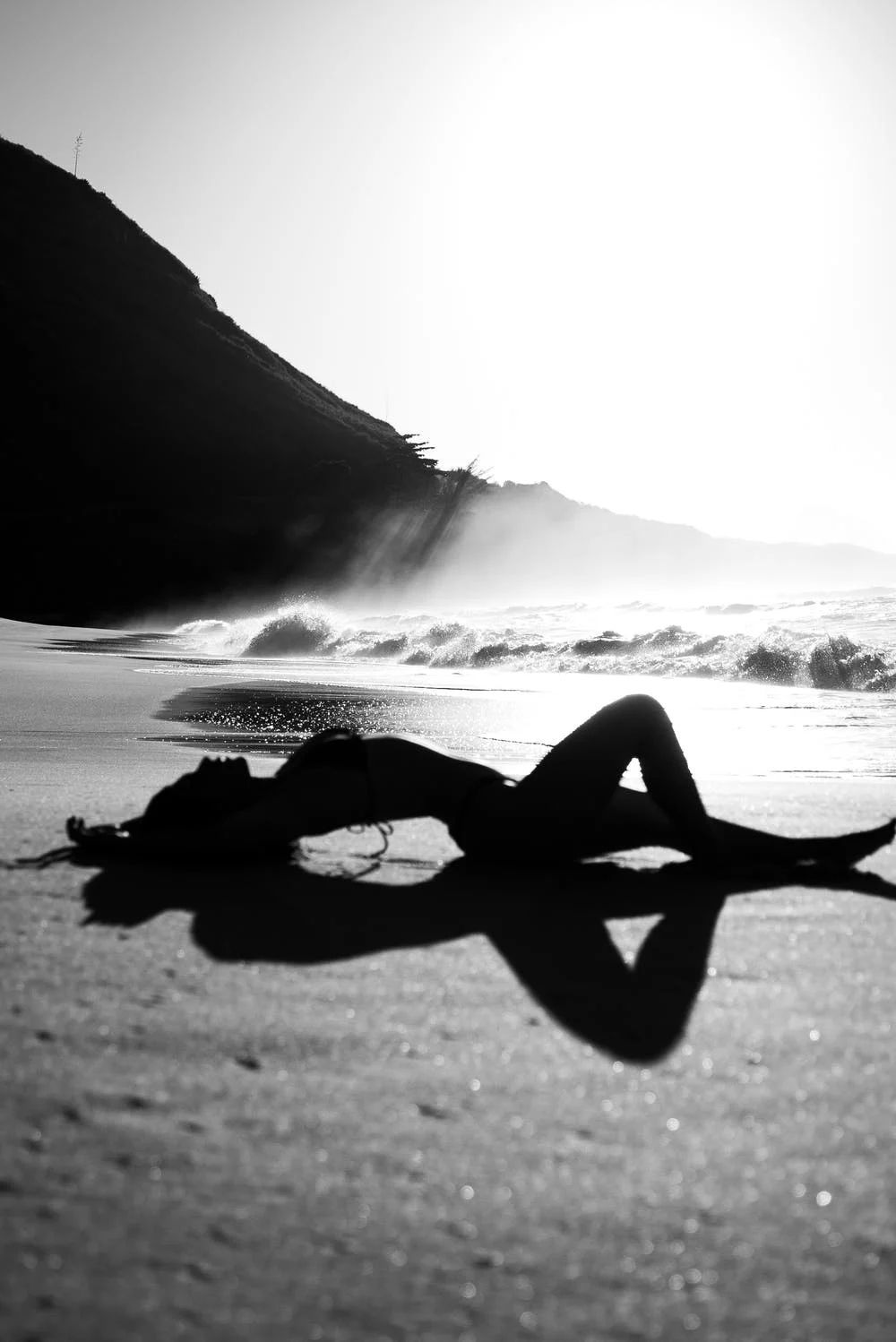 Silhouette of a woman lying on a sunlit beach with crashing waves and a rocky cliff in this black and white photo.