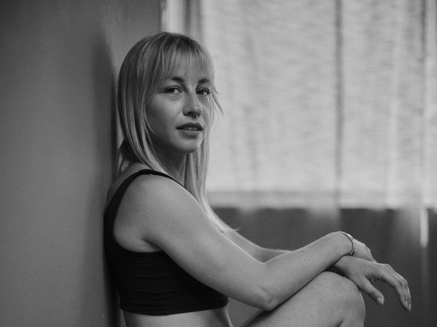 Black and white portrait of a young blonde woman in a crop top leaning against a wall, looking thoughtful.