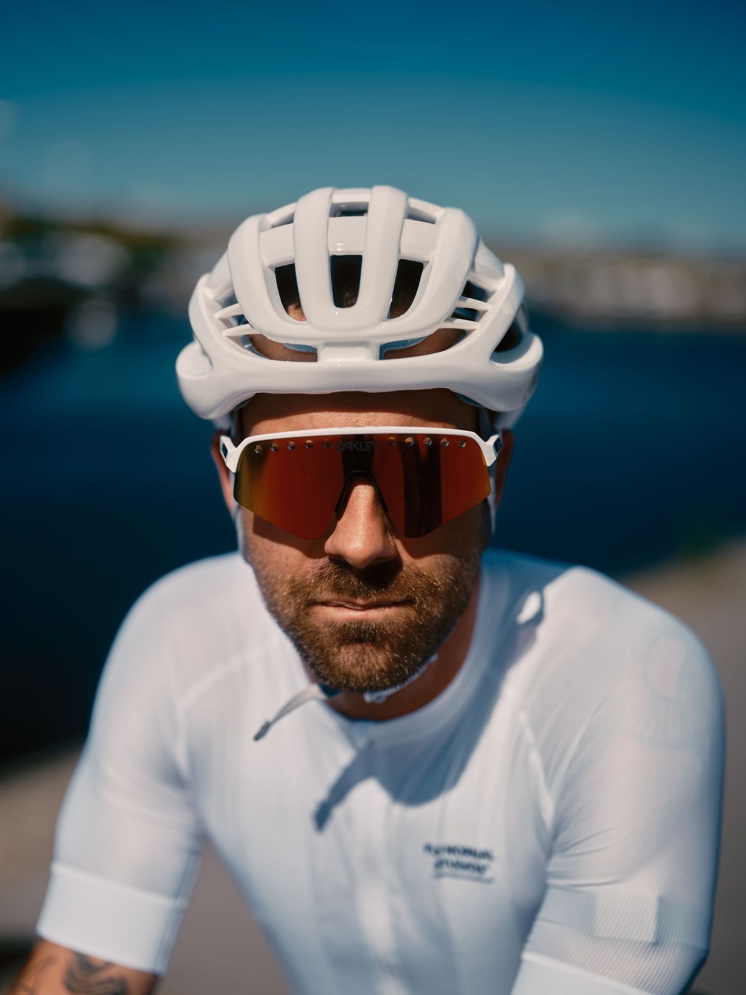 Male cyclist in white helmet and red mirrored sunglasses wearing white jersey, posing outdoors near water on sunny day.