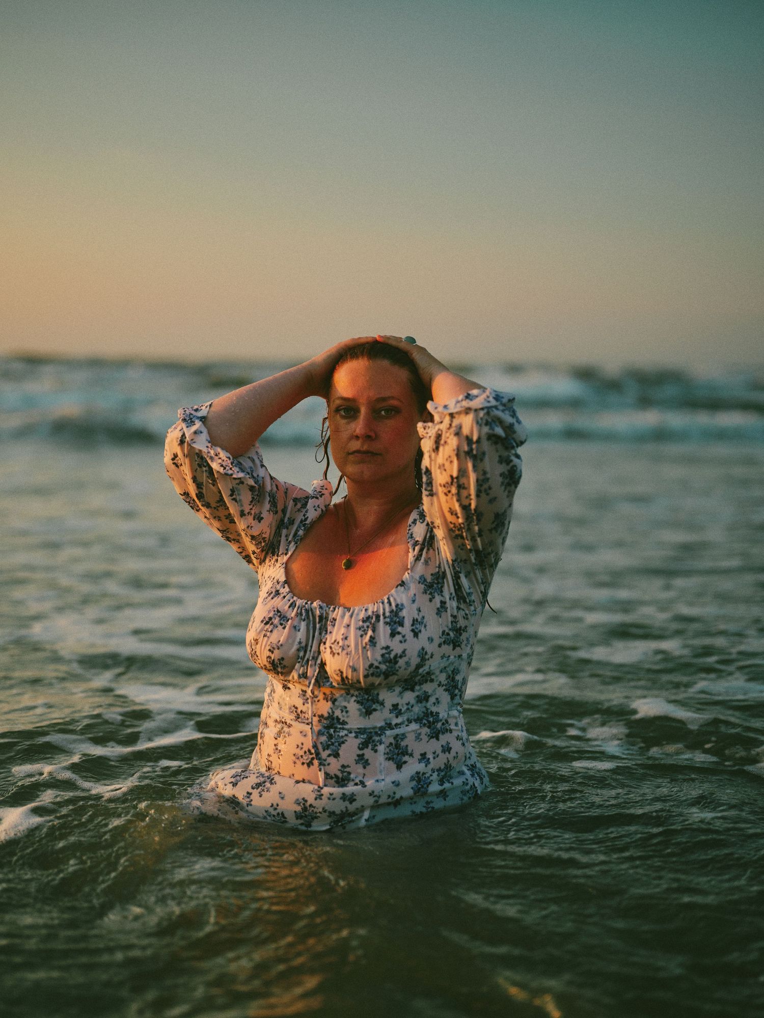 Woman in floral dress standing in ocean waves at sunset, hands in hair, wearing glasses.