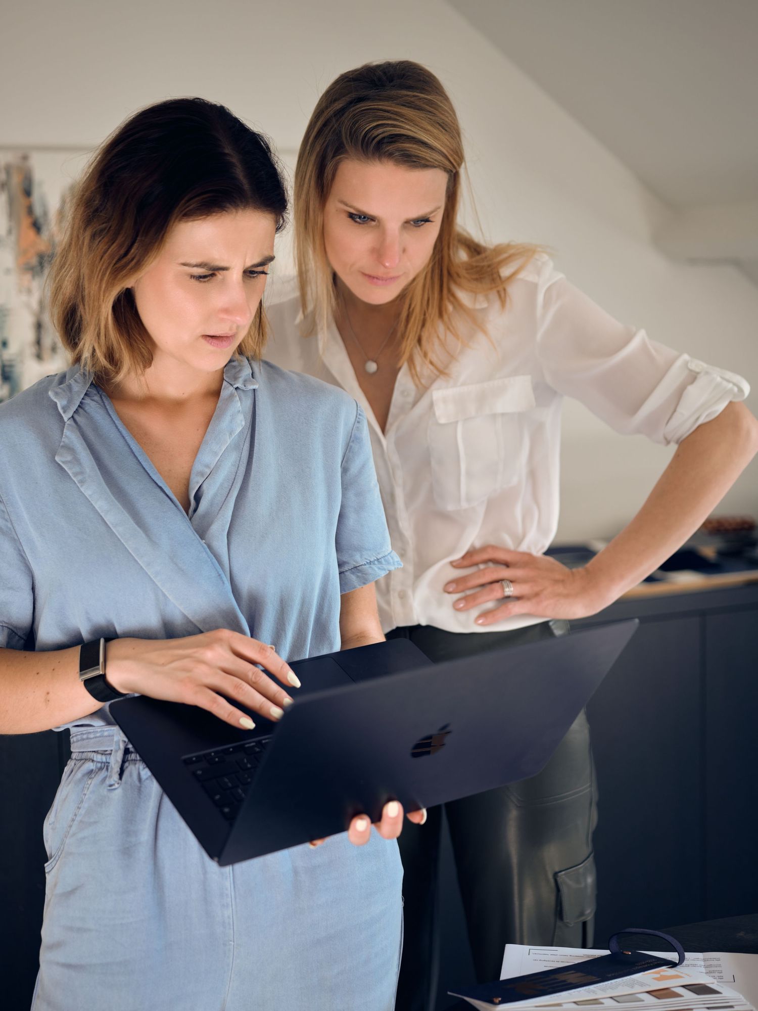 Two women collaborating over a dark MacBook laptop in a modern office setting.