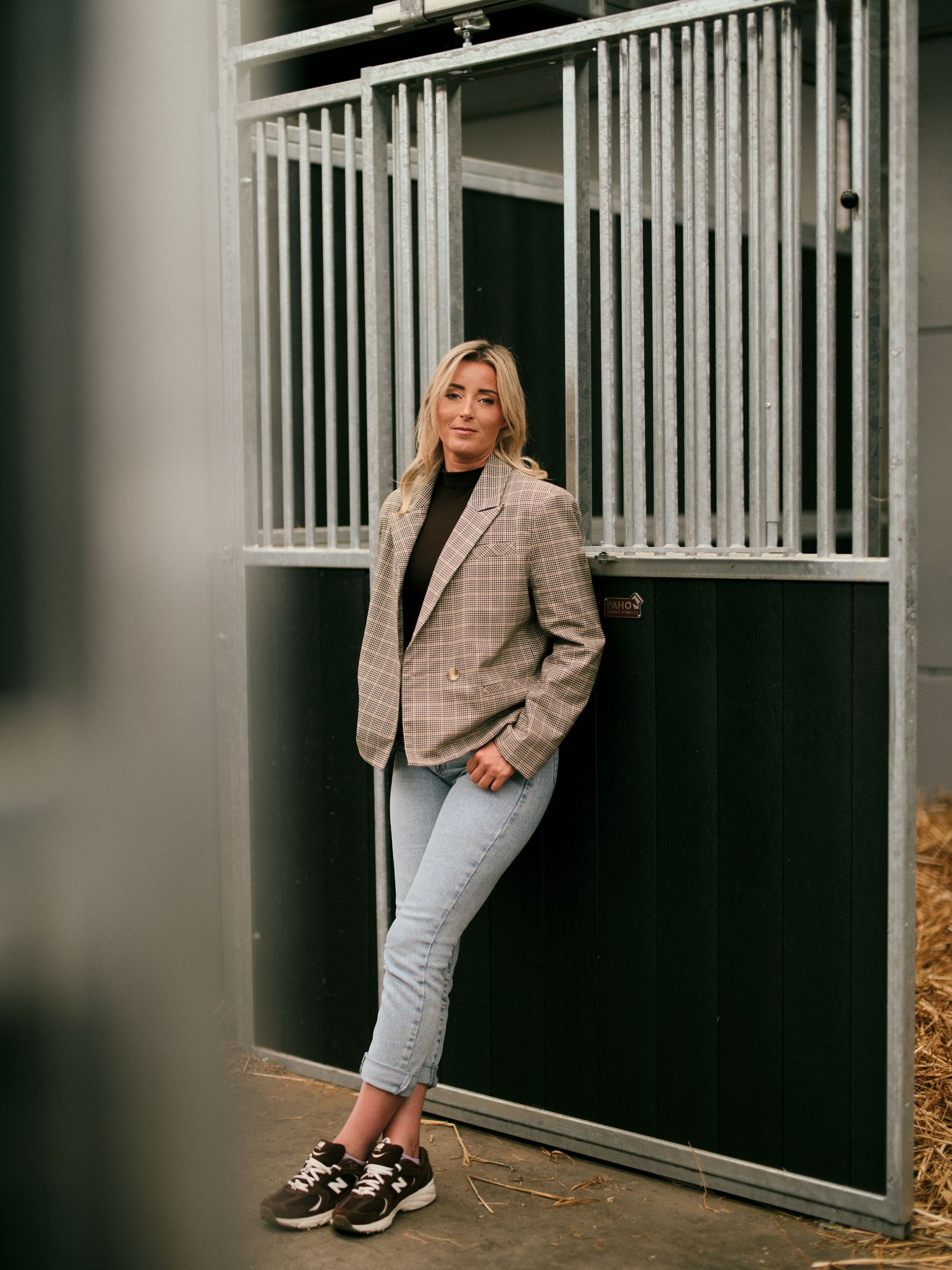 Woman in plaid blazer, black turtleneck, and jeans poses leaning against a horse stable door.