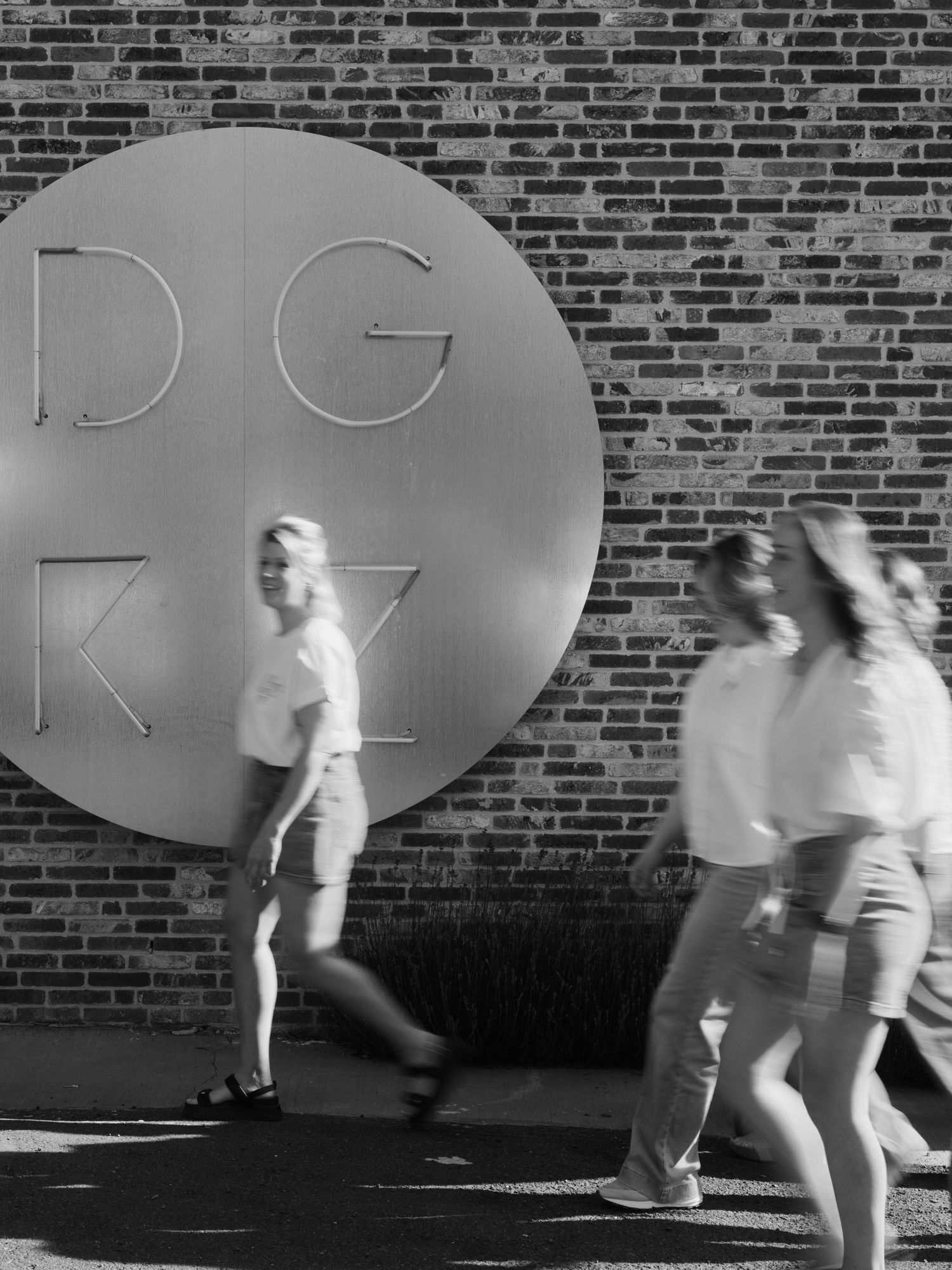 Black and white photo of people walking past a large circular DGKV sign on a brick wall.