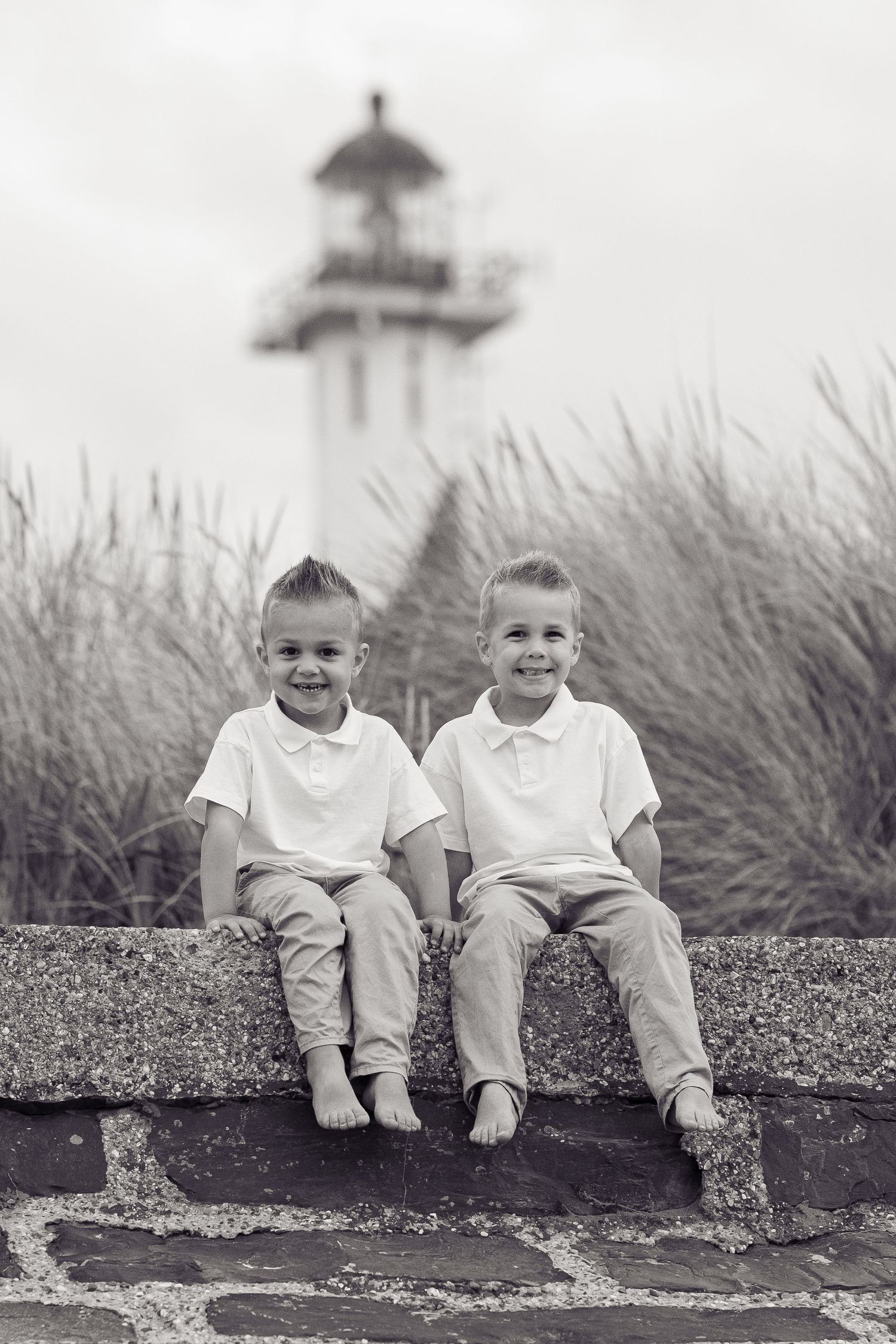 Black and white photo of children sitting by lighthouse with coastal grasses.