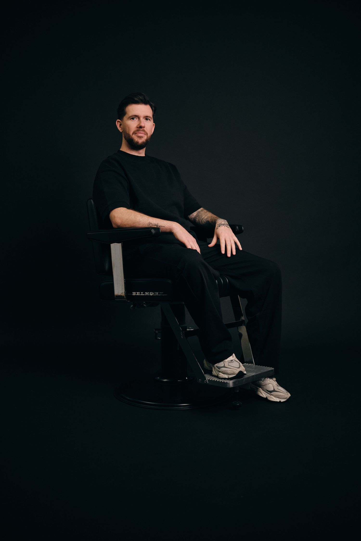 Person in all black attire sits casually on chair against dark background in moody studio portrait.