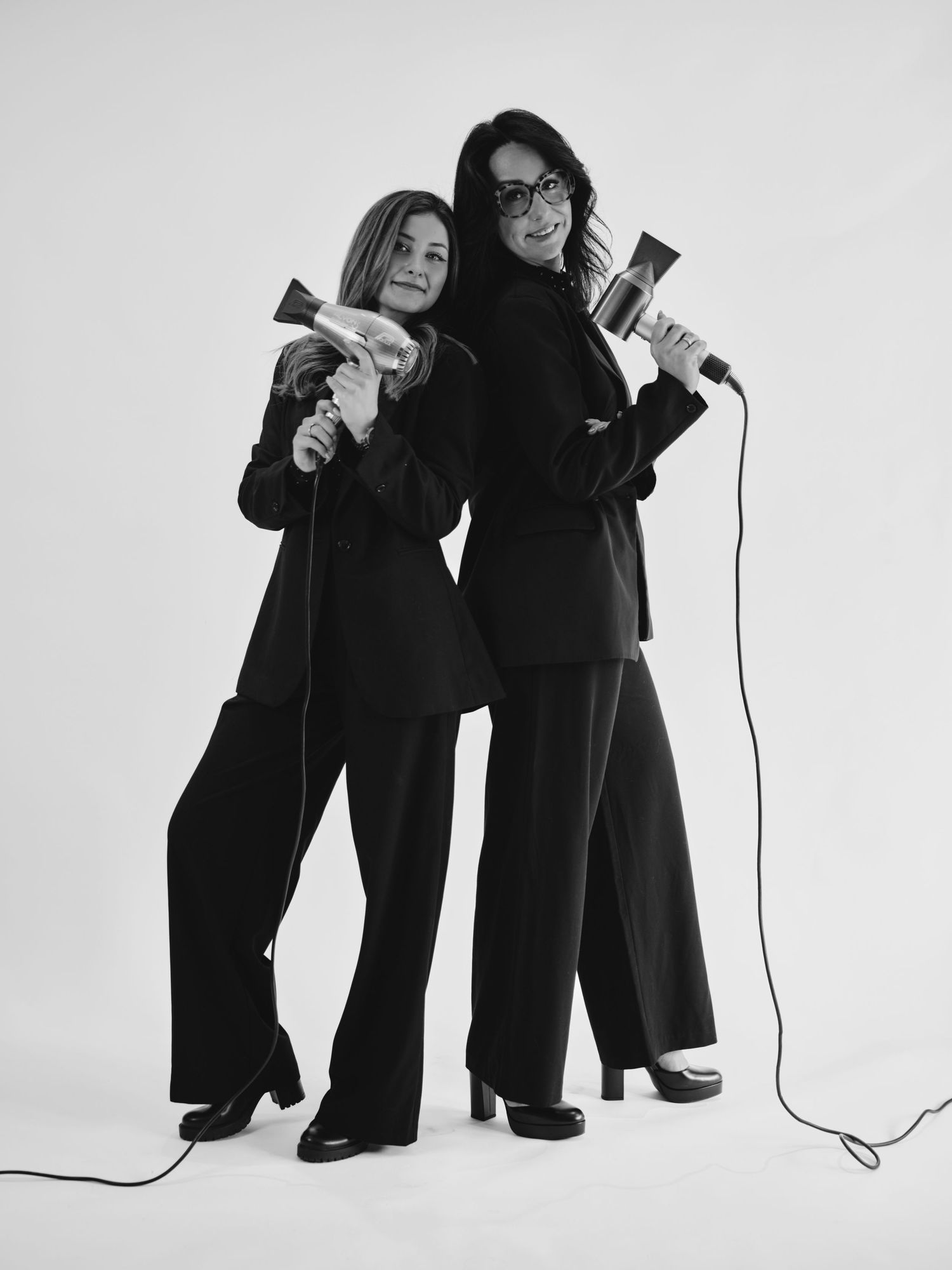 Two women in black suits holding hair dryers back-to-back in a black and white studio photo.