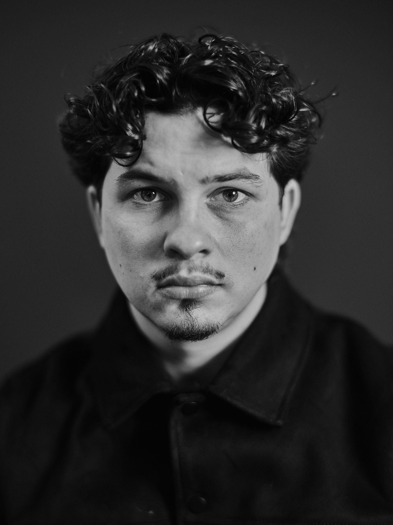 Black and white portrait of a young man with curly hair and a goatee wearing a dark jacket.
