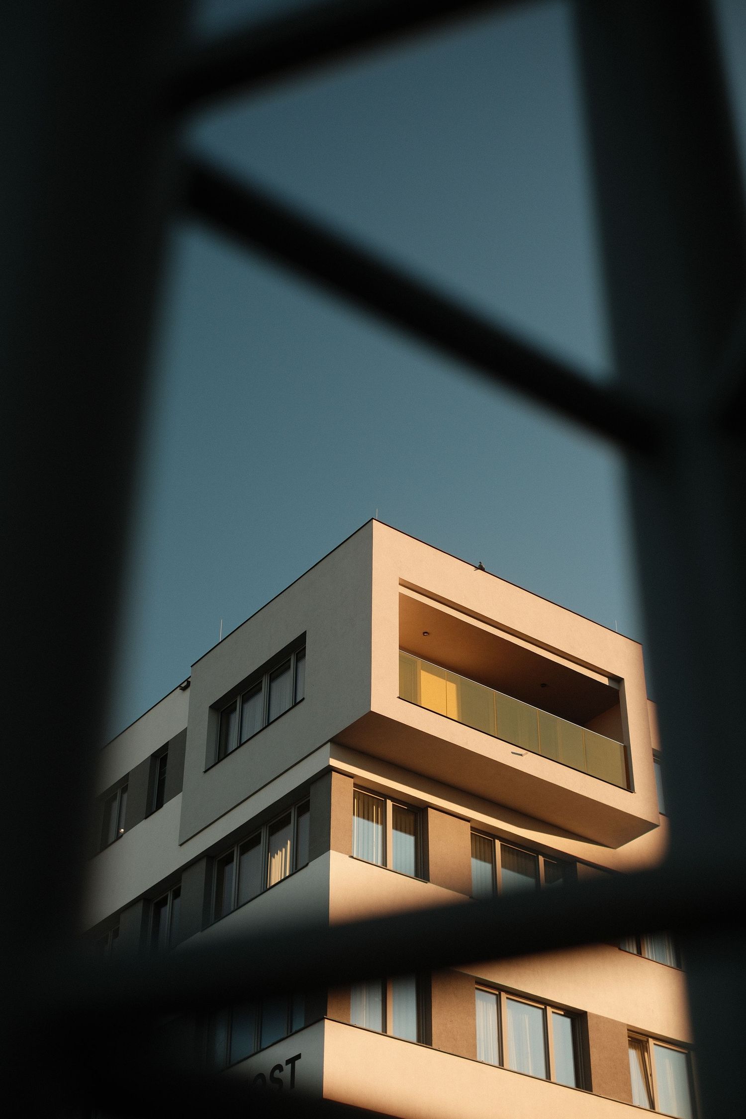 Modern apartment building photographed through blurred foreground bars, bathed in warm golden sunlight against a clear blue sky.