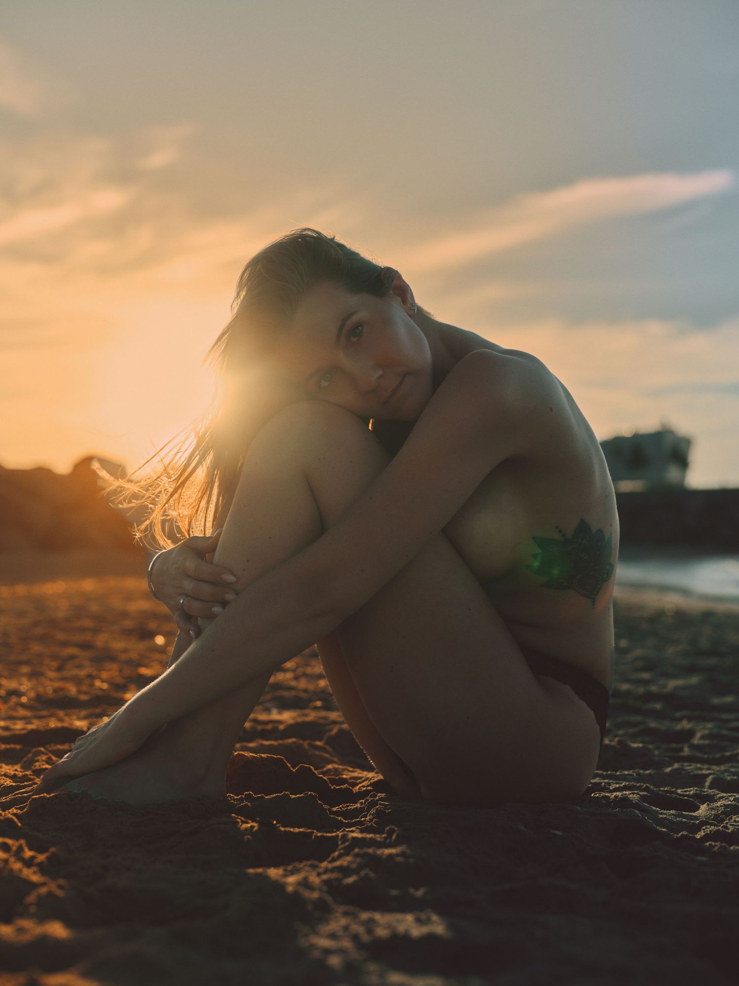 Woman with tattoo sitting on sandy beach at golden sunset, hugging knees and smiling warmly.