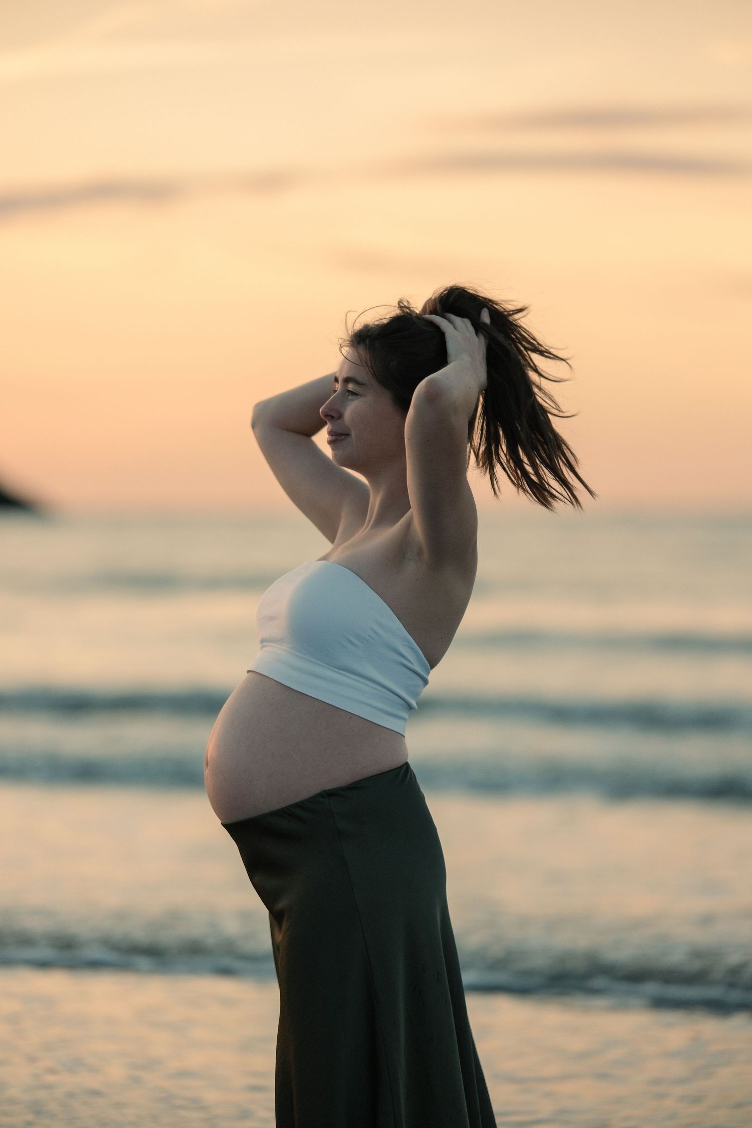 Silhouetted maternity portraits against sunset sky at beach with waves in background.