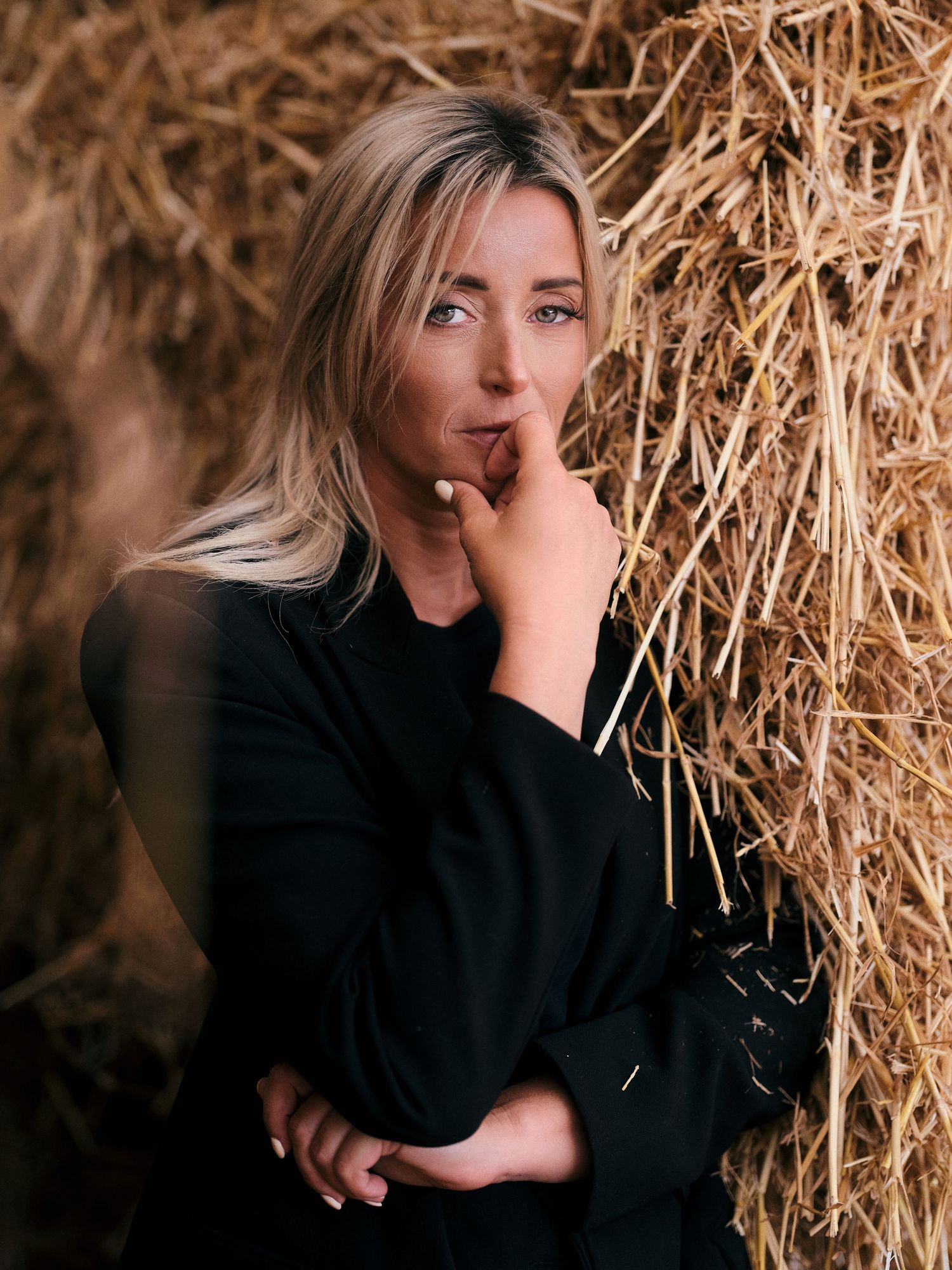 Blonde woman in black outfit posing thoughtfully against rustic hay bales in an outdoor farm setting.