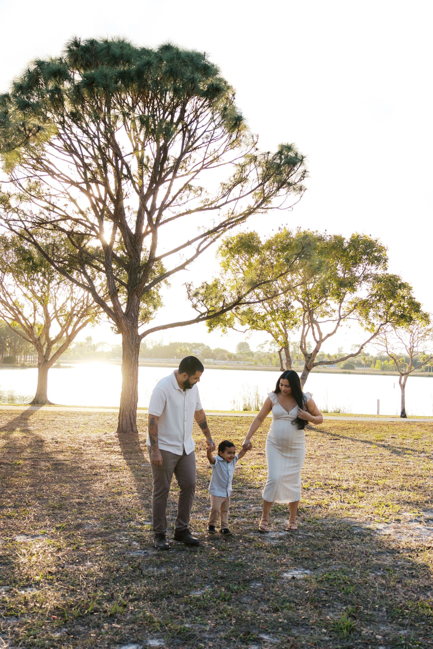 Family of three walking hand-in-hand in a sunlit park near a lake, parents in white outfits with a toddler between them.