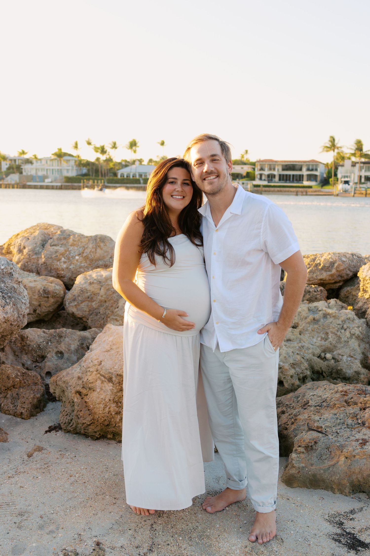 Smiling pregnant couple posing together on rocky beach at golden hour in white outfits.