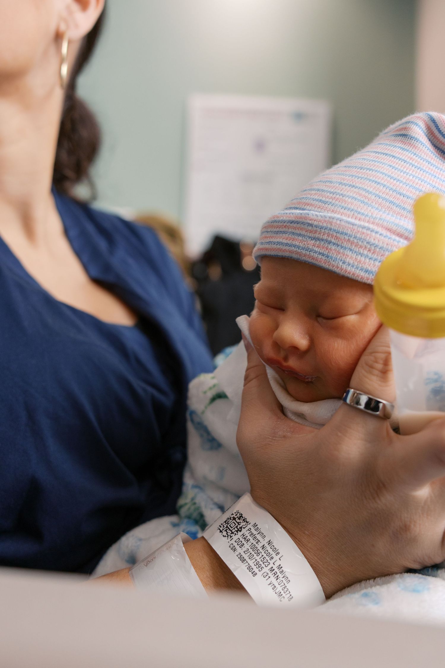Newborn baby in striped hat held gently by parent with hospital ID bracelet visible in maternity ward.