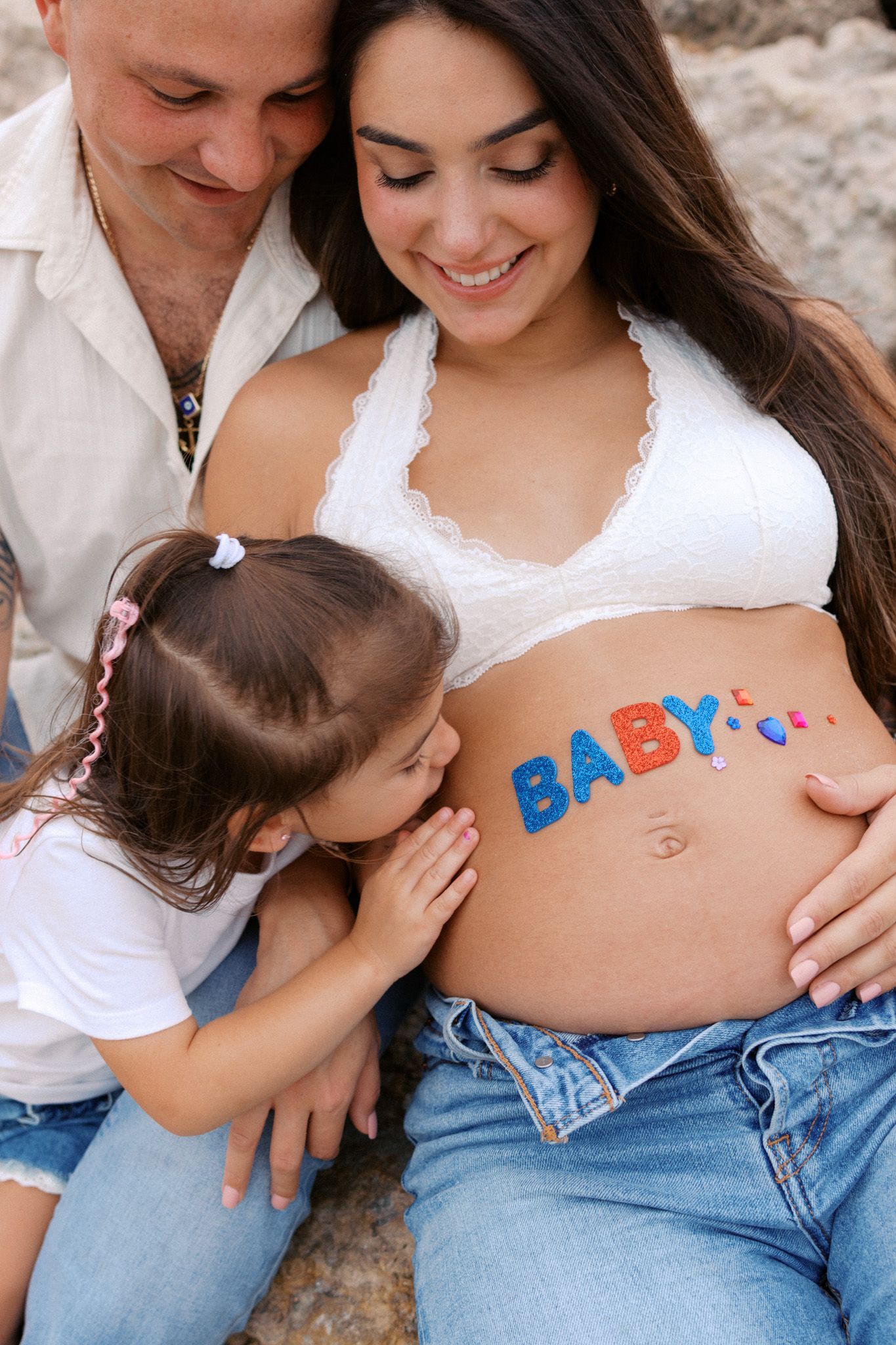 A pregnant woman with BABY painted on her belly, kissed by her daughter, while her husband smiles beside them outdoors.