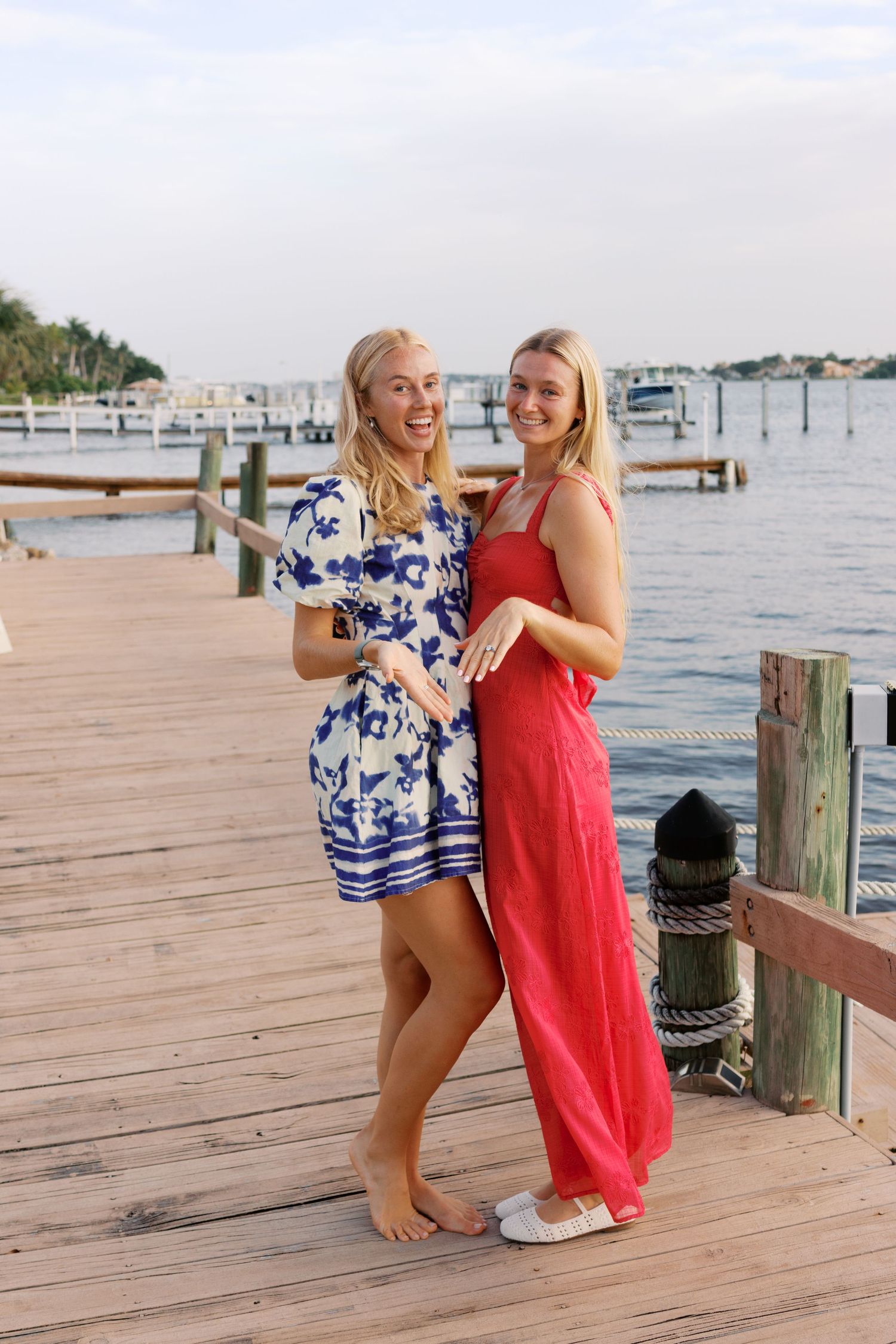 Two blonde women in summer dresses pose together on a wooden marina dock with water and boats in the background.