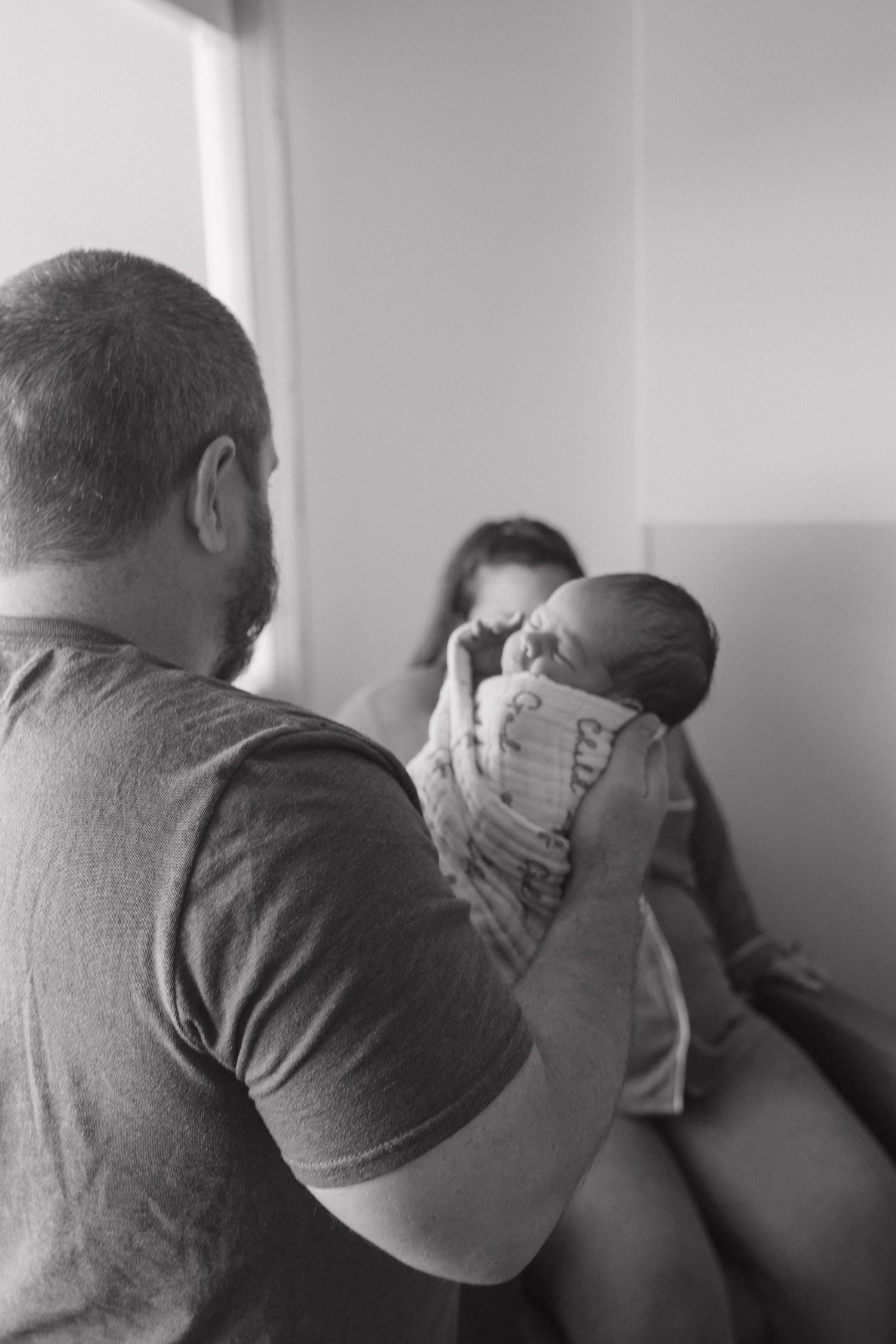 Black and white photo of a father holding a swaddled newborn baby while mother watches tenderly in background.