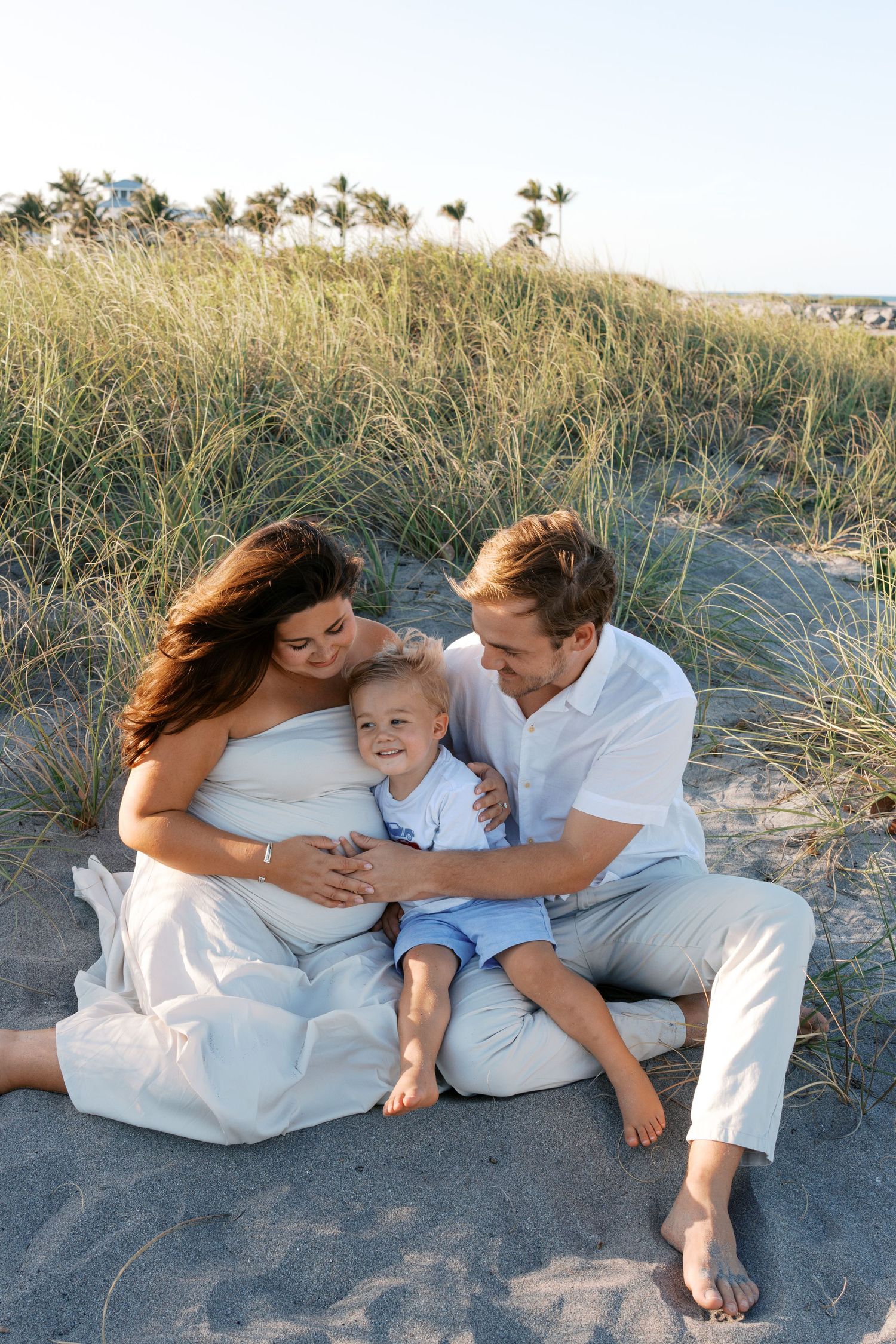 Pregnant woman and man sit on beach sand with toddler boy, surrounded by dune grass in a family maternity photo.