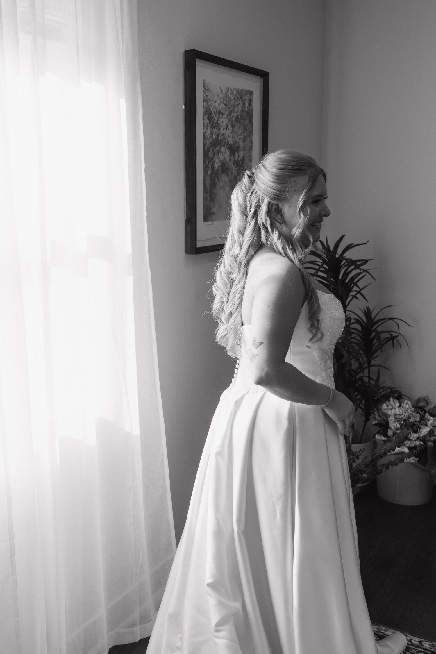 Elegant bride in white satin gown stands by window in black and white wedding portrait.