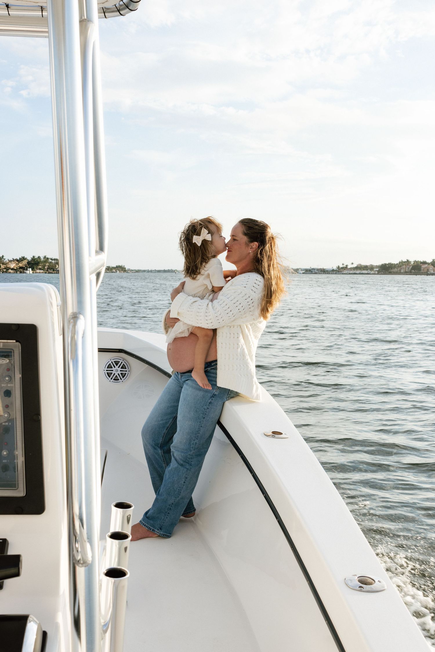 A mother holds her young daughter on a white boat deck, sharing a tender kiss against a serene waterway backdrop.