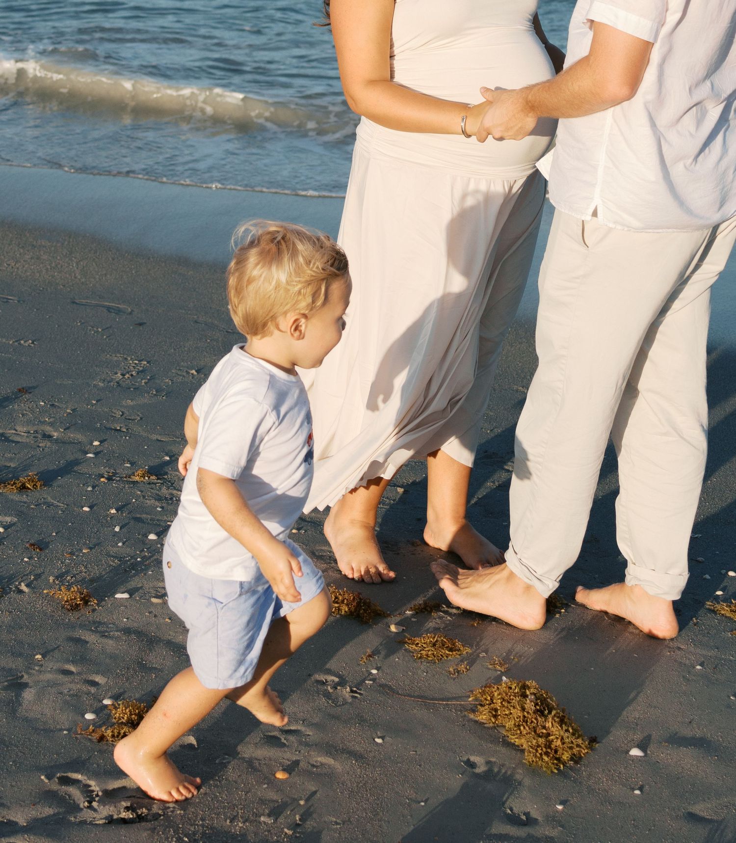 A toddler walks barefoot on a sandy beach beside two adults in light clothing, with gentle ocean waves in the background.