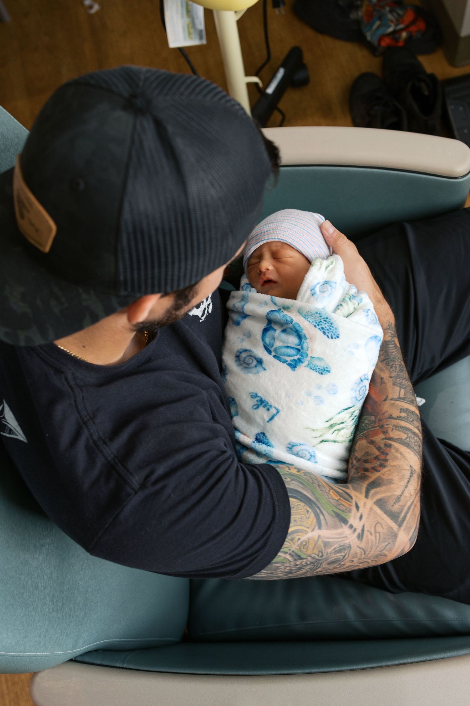 Tattooed father in black cap holding swaddled newborn baby in hospital chair.