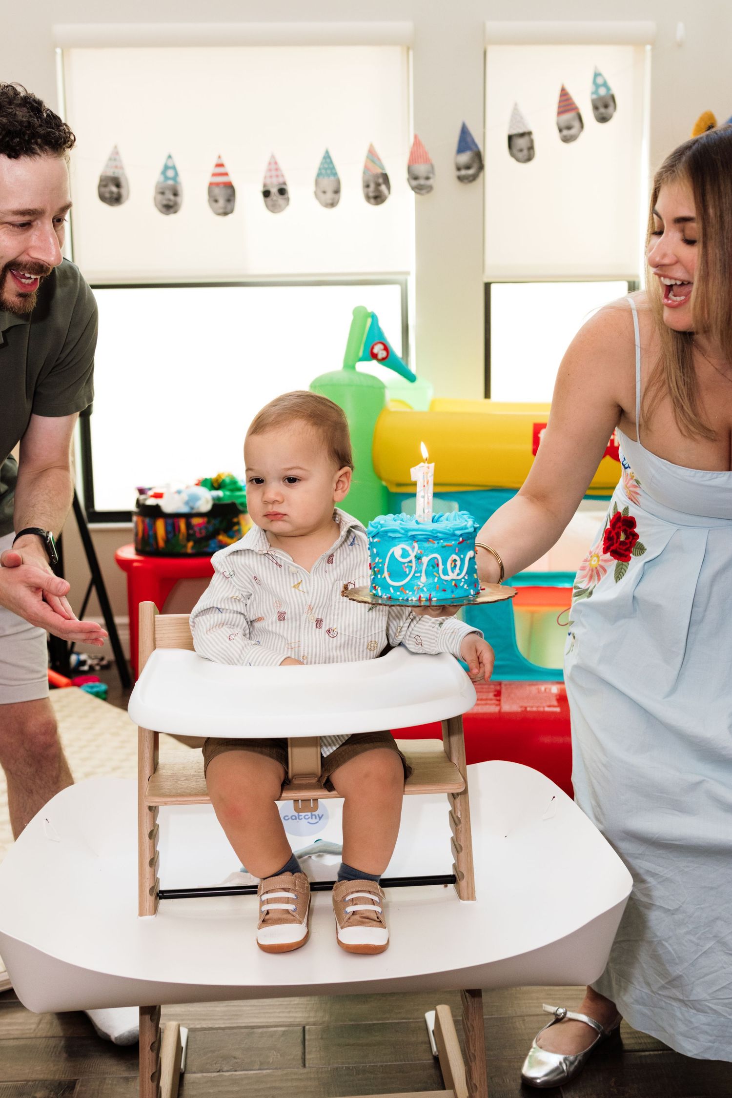 Parents celebrating baby's first birthday with a blue 'One' cake in a colorful playroom.