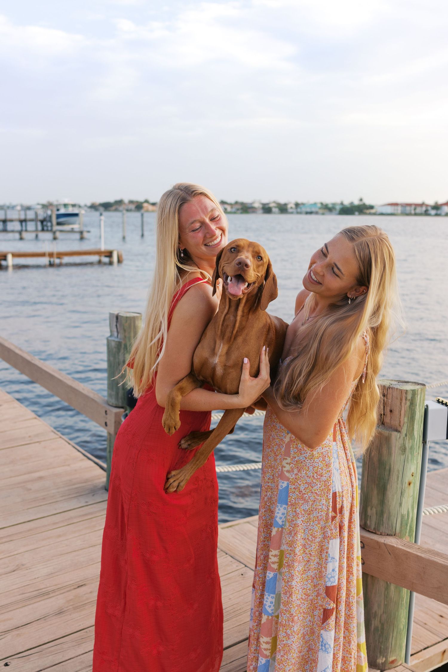 Two blonde women in summer dresses laugh with a happy brown dog on a waterfront dock at sunset.