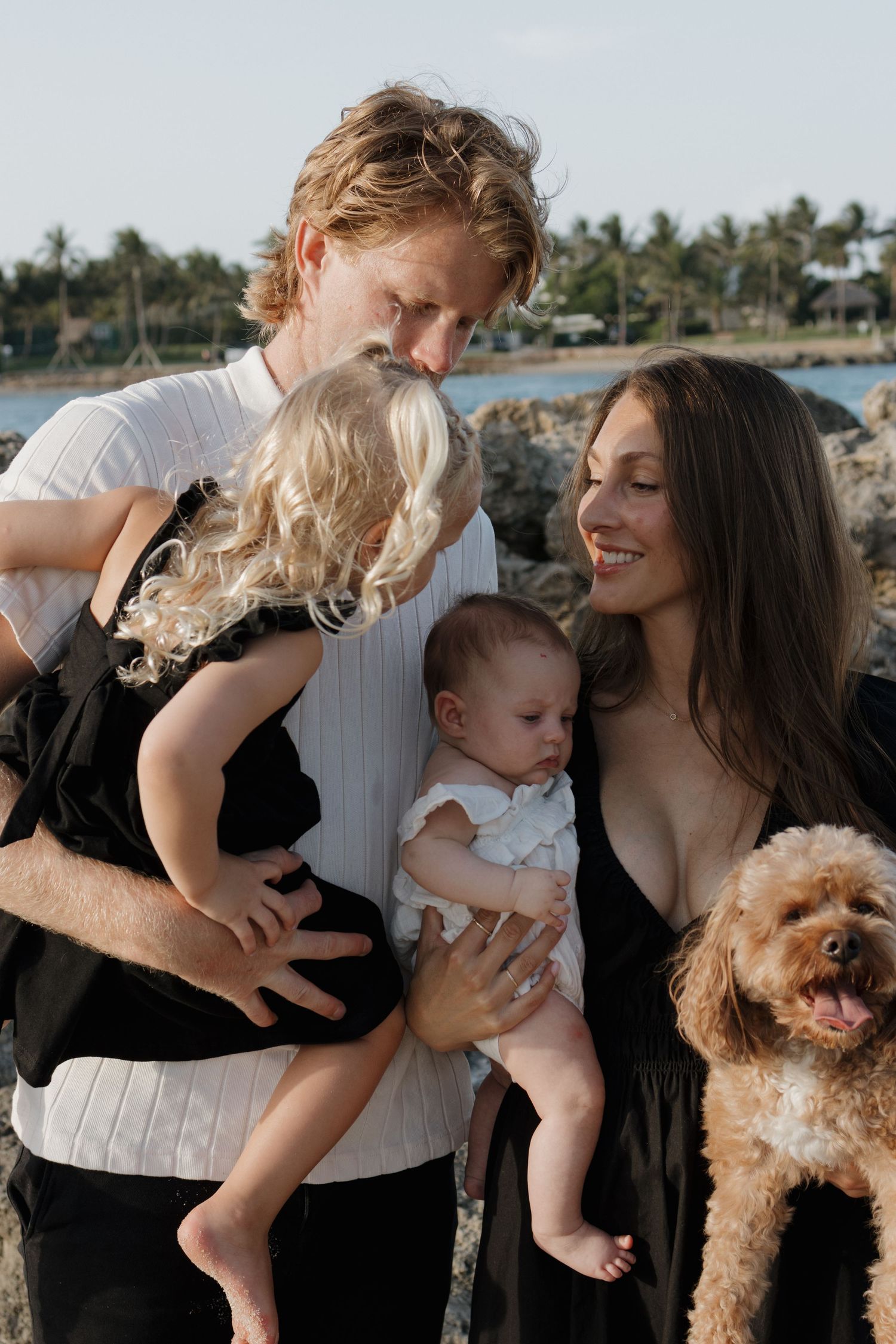 A family poses together with their small dog on a tropical beach with palm trees in the background.