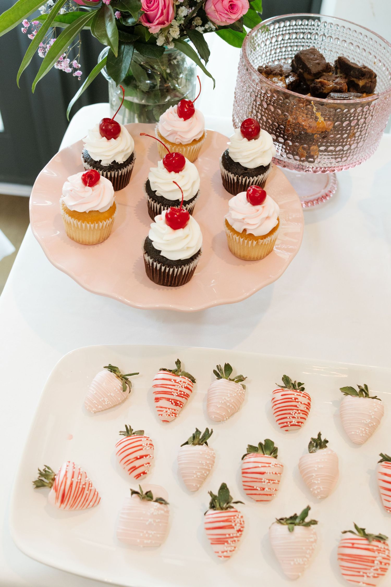Pink cake stand with cherry-topped cupcakes beside white chocolate-drizzled strawberries on a dessert table.