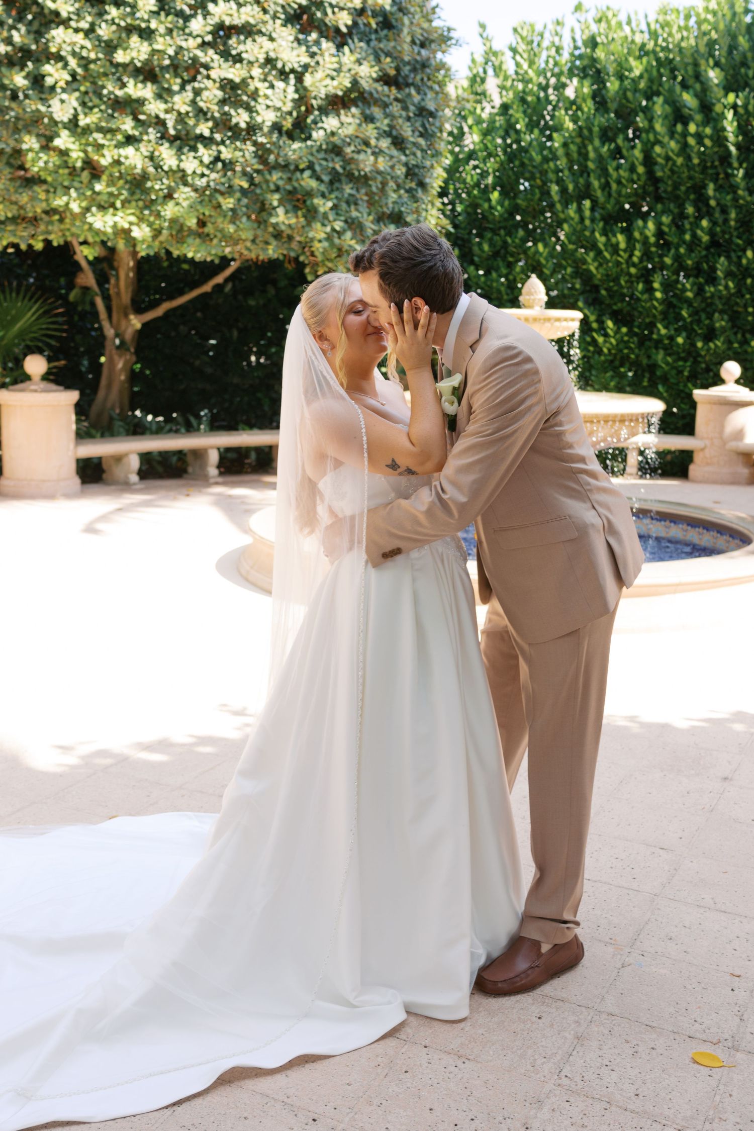 Bride and groom sharing first kiss in white gown and tan suit beside garden fountain on sunny wedding day.