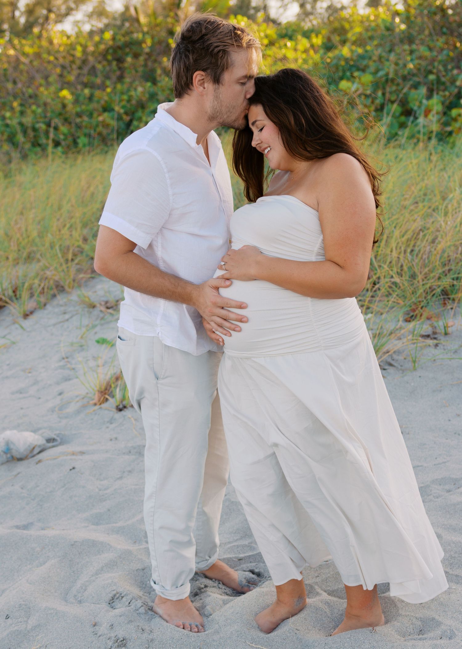 Expecting couple in white outfits share a tender moment on sandy beach during maternity photography session.