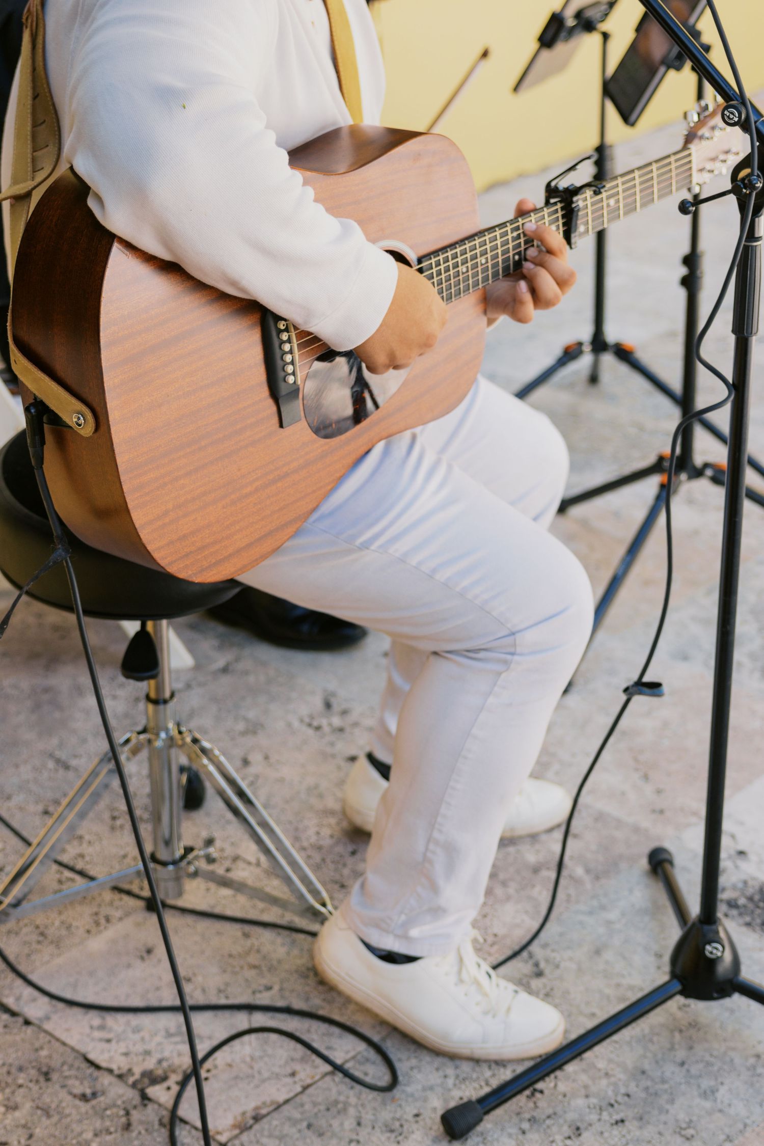 Person in white outfit sitting on stool playing acoustic guitar outdoors near microphone stands.