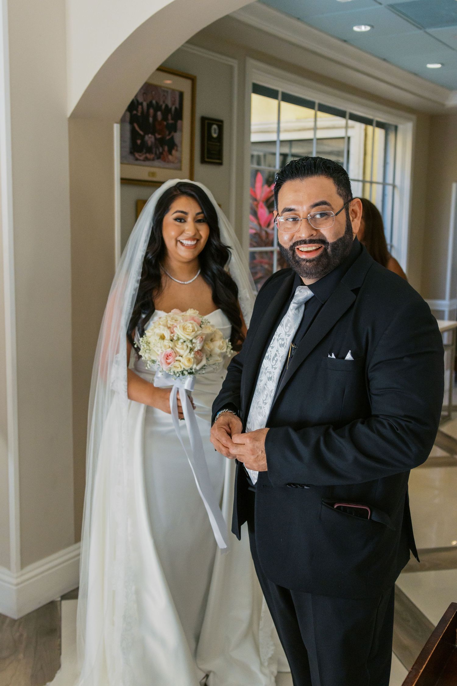 Bride in white gown holding bouquet stands with suited man in glasses, smiling before wedding ceremony.