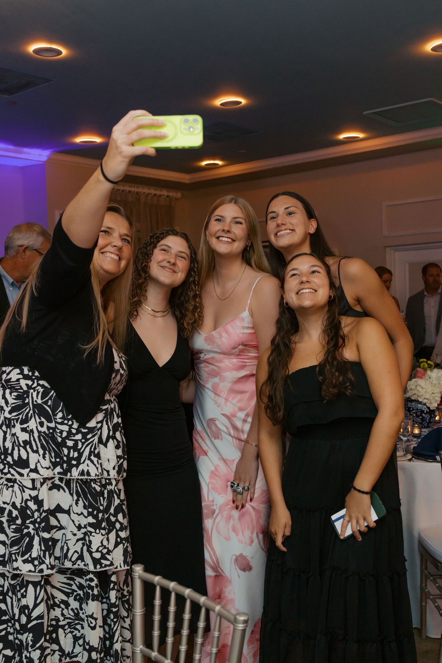 Five young women in formal dresses pose together for a selfie at an elegant indoor event or wedding reception.