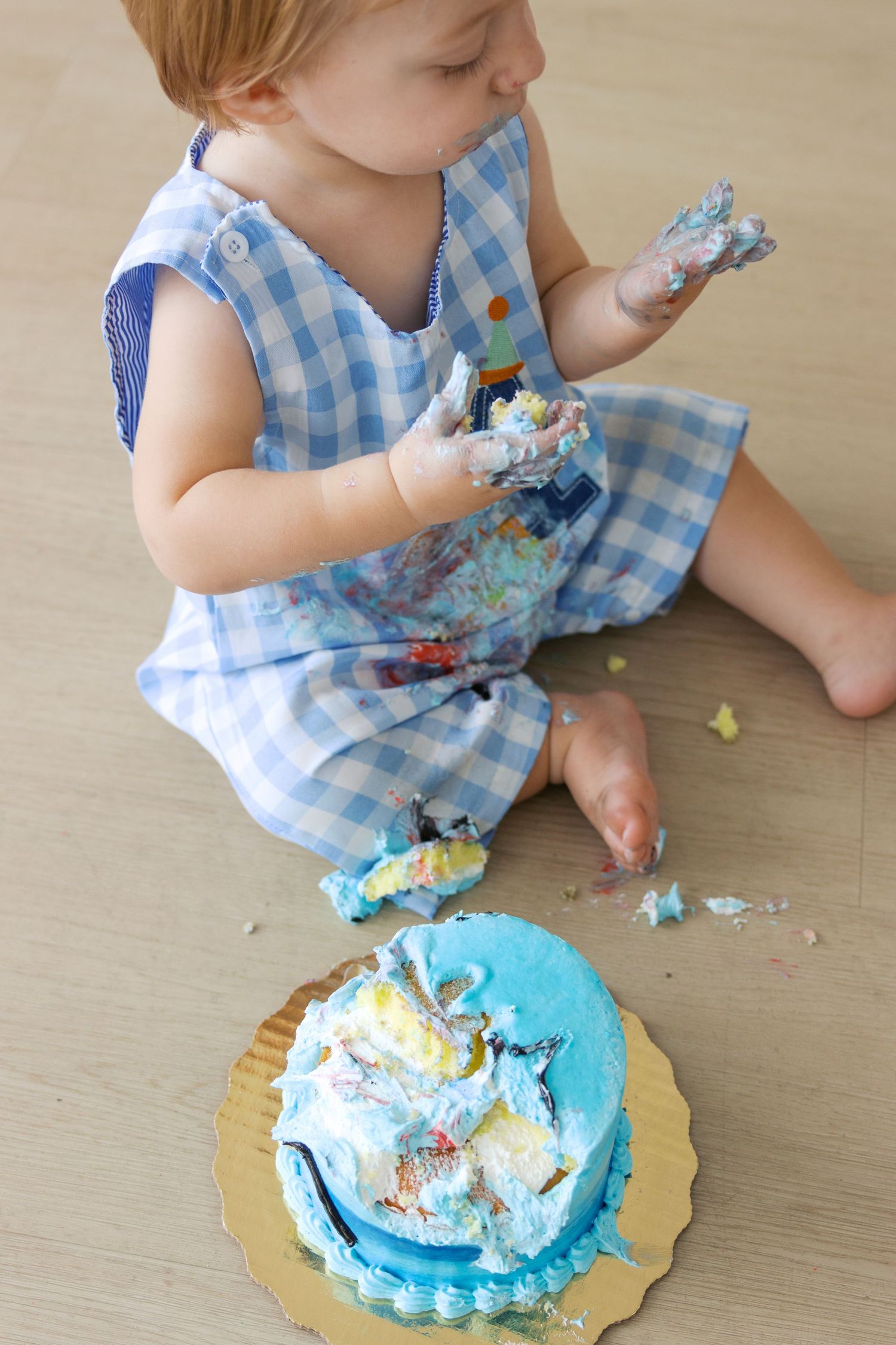 Baby in blue gingham dress smashing a blue frosted first birthday cake, with frosting on hands and floor.
