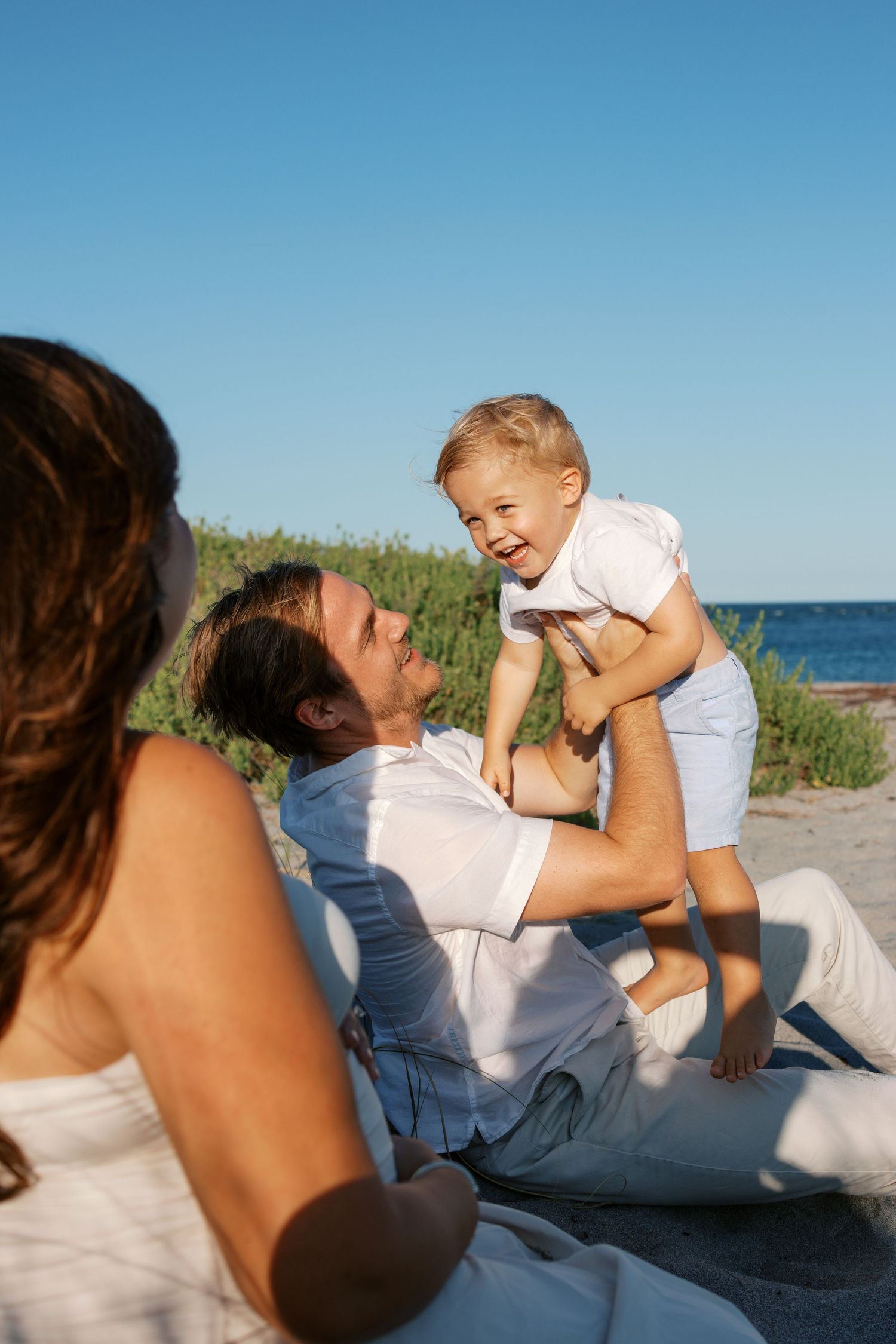 Happy family on beach, father lifting laughing toddler while mother watches, sunny day with ocean in background.