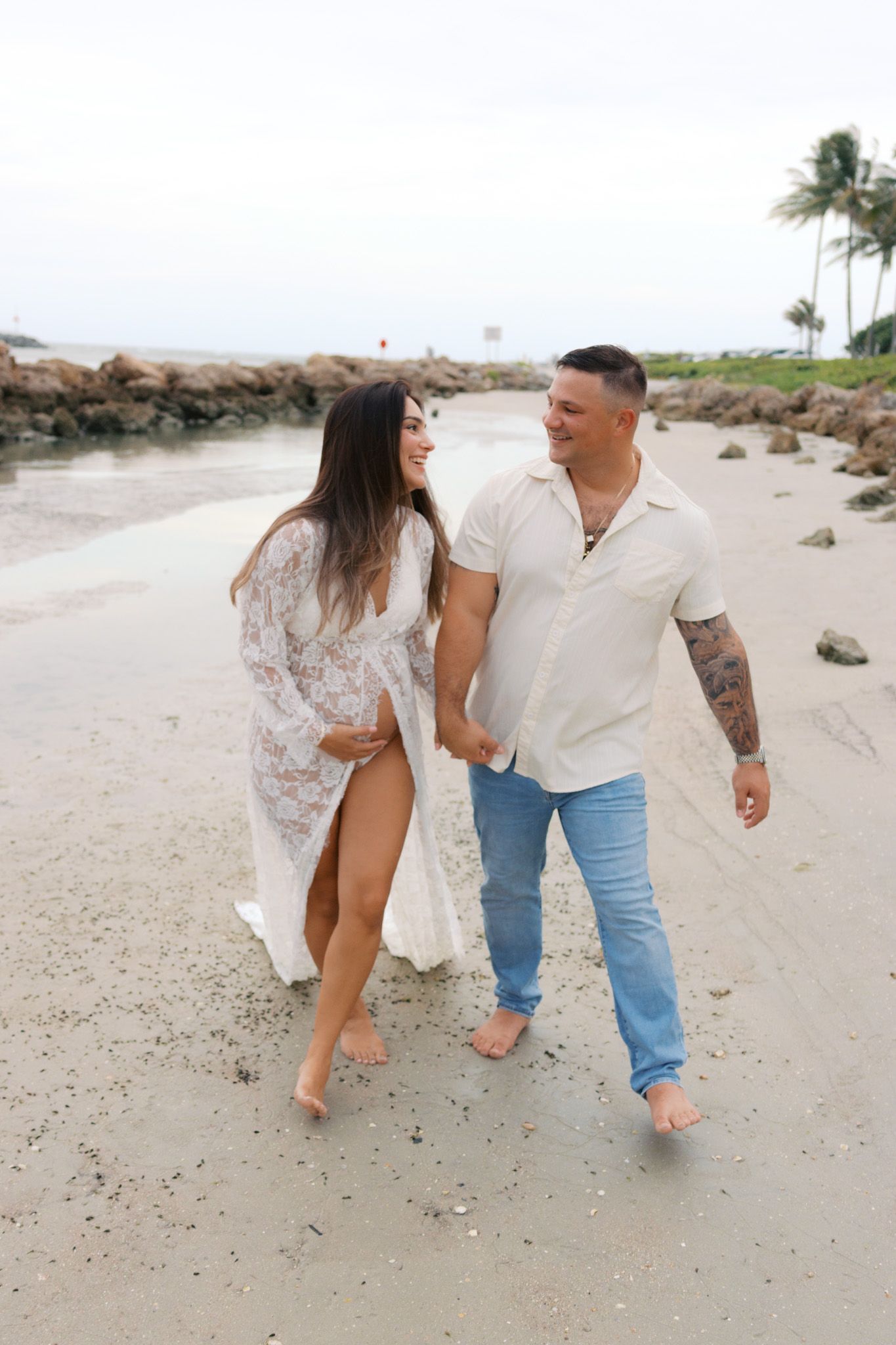 Couple walking barefoot on sandy beach, woman in white lace dress, man in cream shirt and jeans, holding hands and smiling.