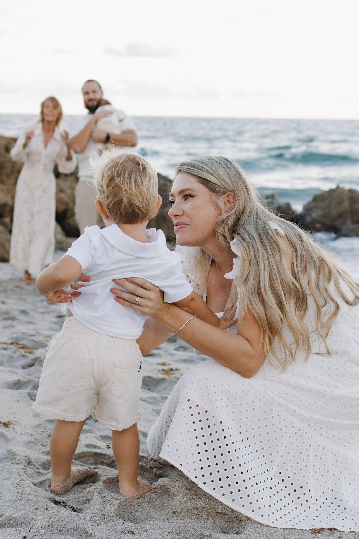 A blonde woman in a white dress crouches to embrace a toddler on a sandy beach, with a family in the background.
