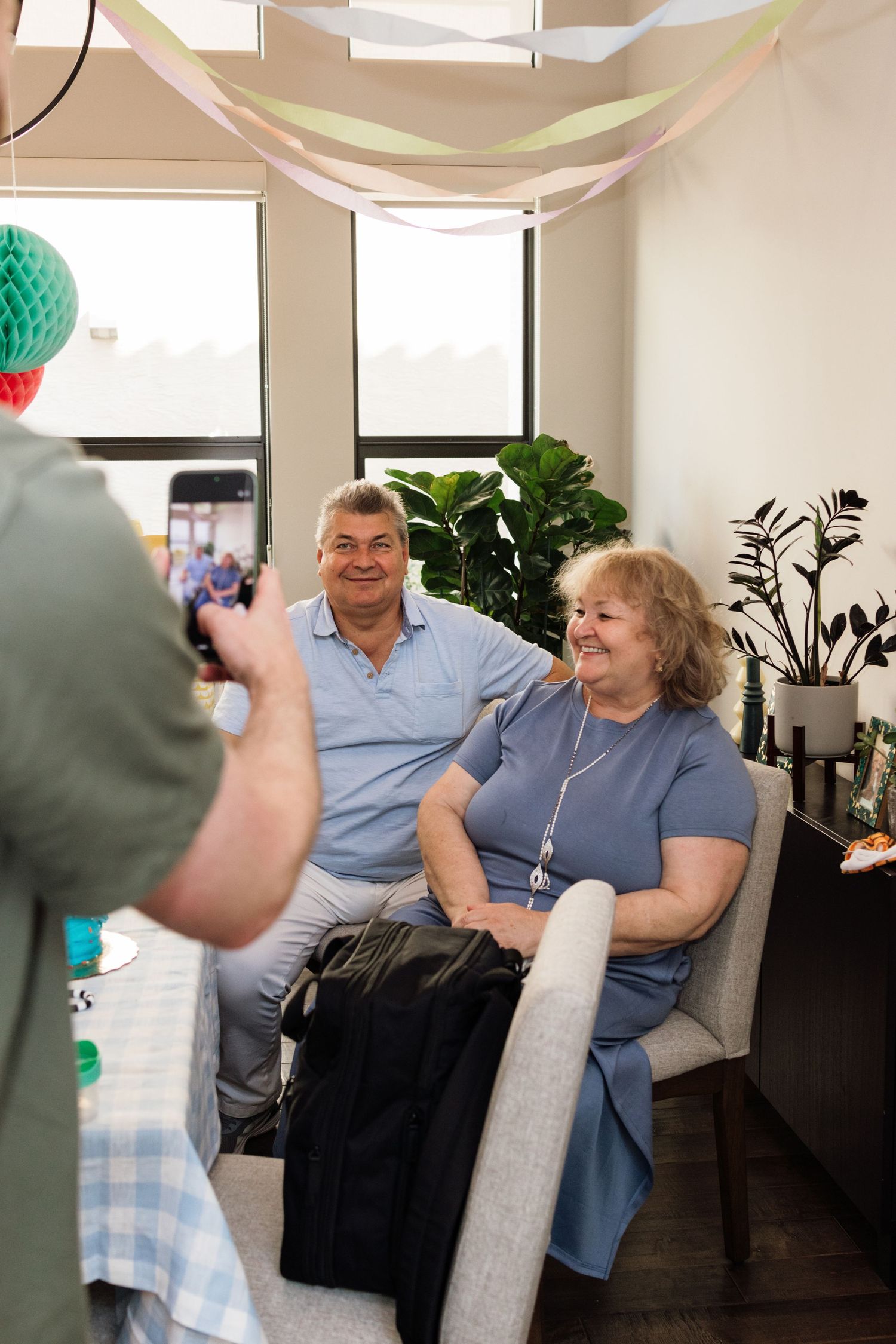 Elderly couple smiling while being photographed at a festive indoor gathering with colorful decorations.