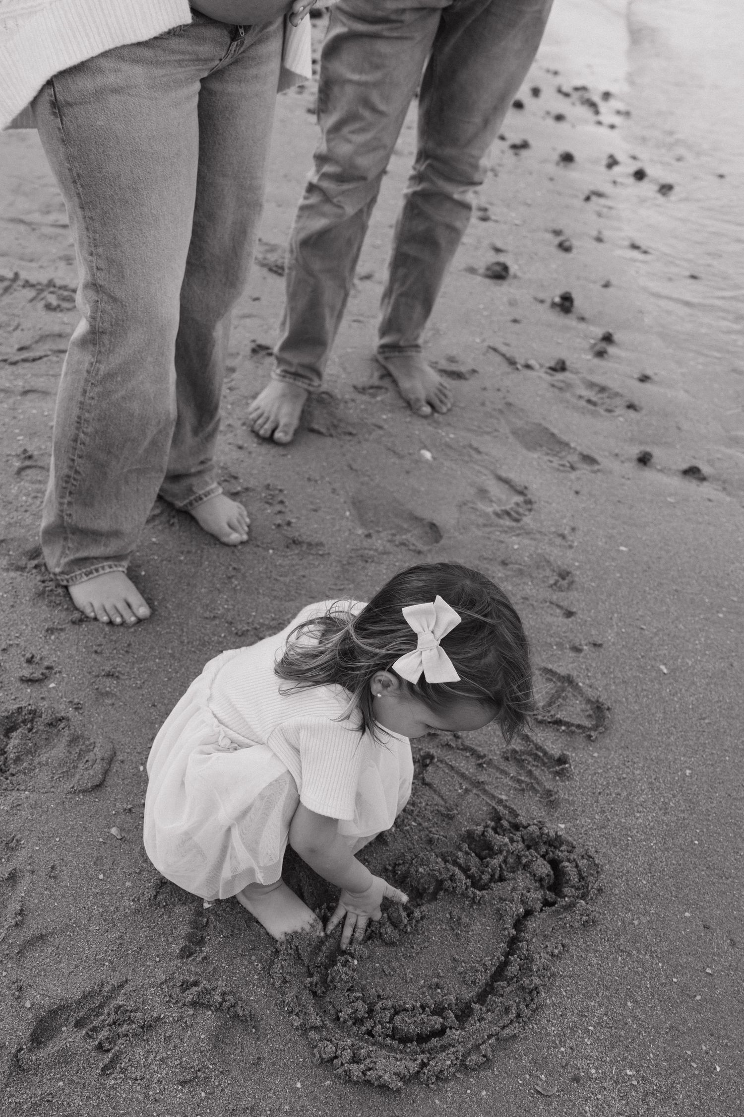 A young girl with a bow draws a heart in beach sand while parents stand nearby, in black and white.