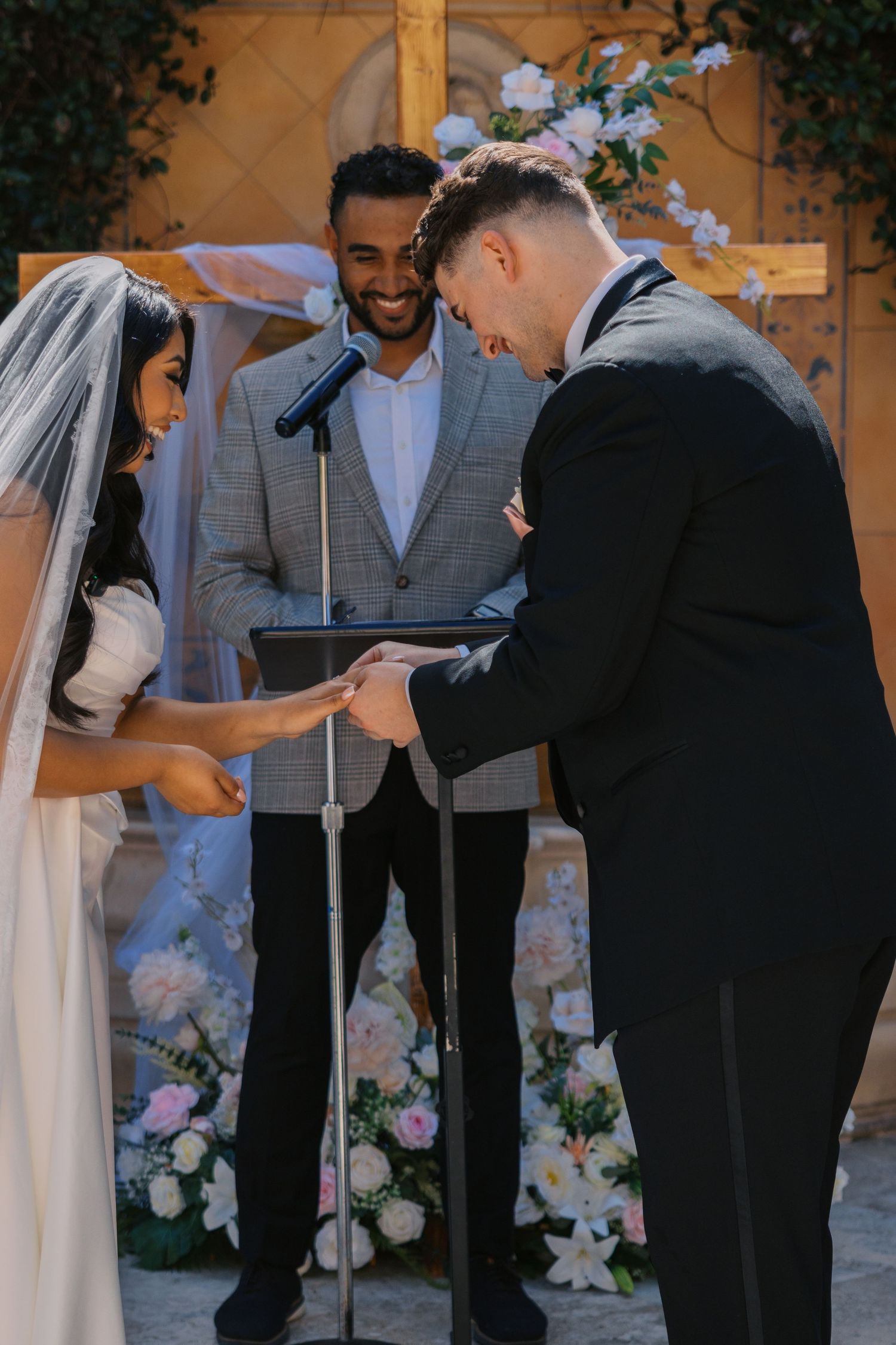 Bride and groom exchanging rings at outdoor wedding ceremony with smiling officiant standing before floral arch.