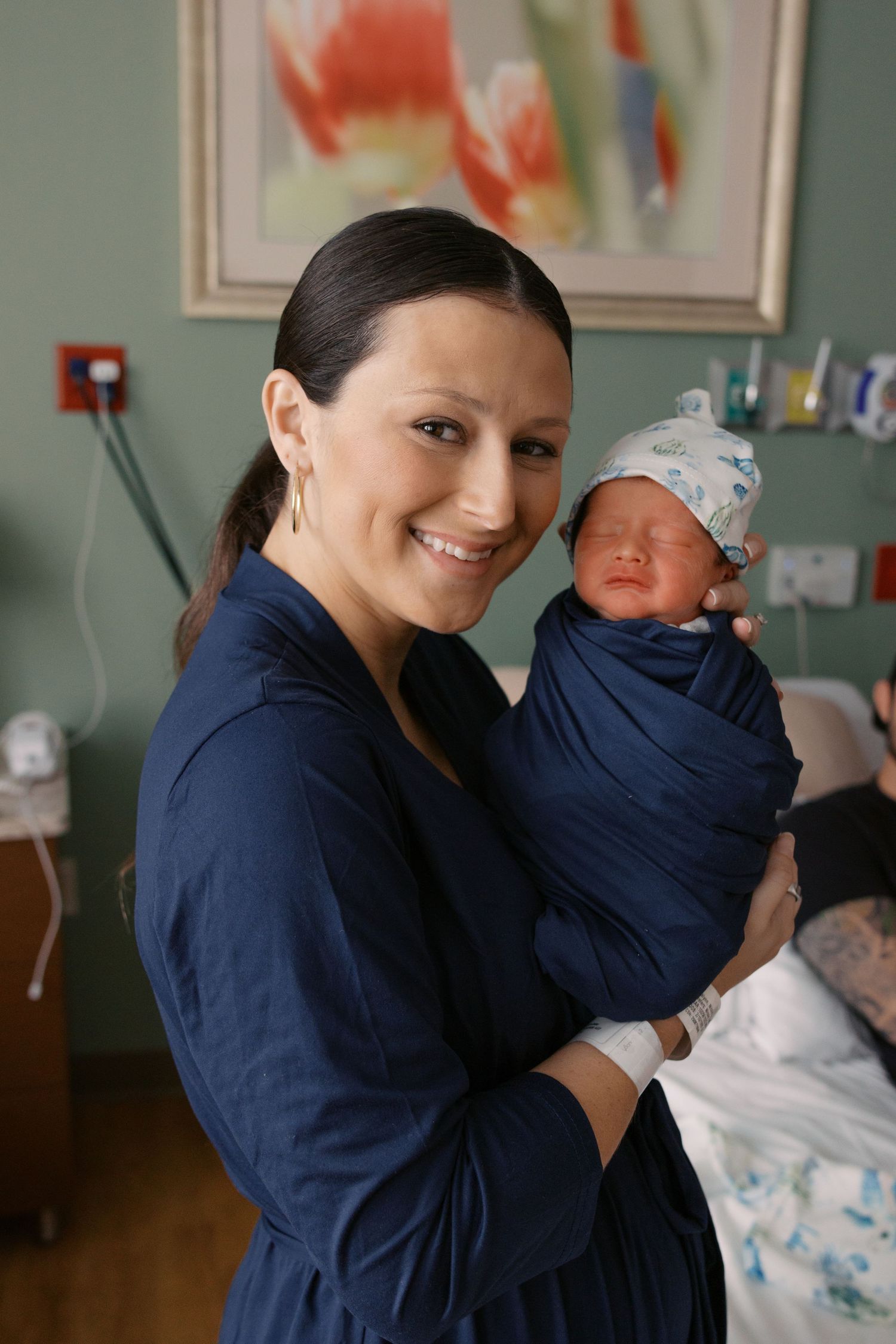 Smiling mother in navy robe holding swaddled newborn baby in hospital room after giving birth.