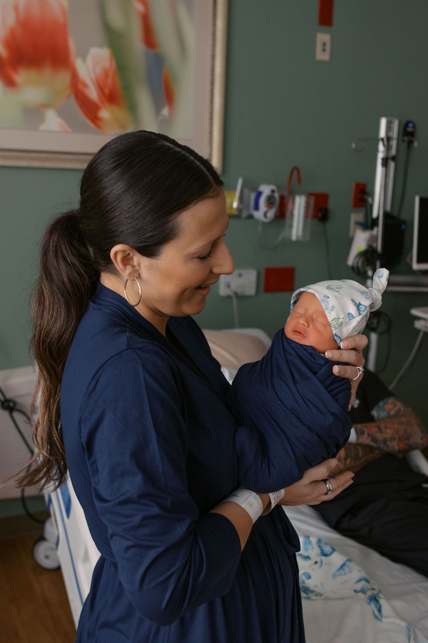 New mother in blue hospital gown smiling while holding her swaddled newborn baby in a maternity ward room.