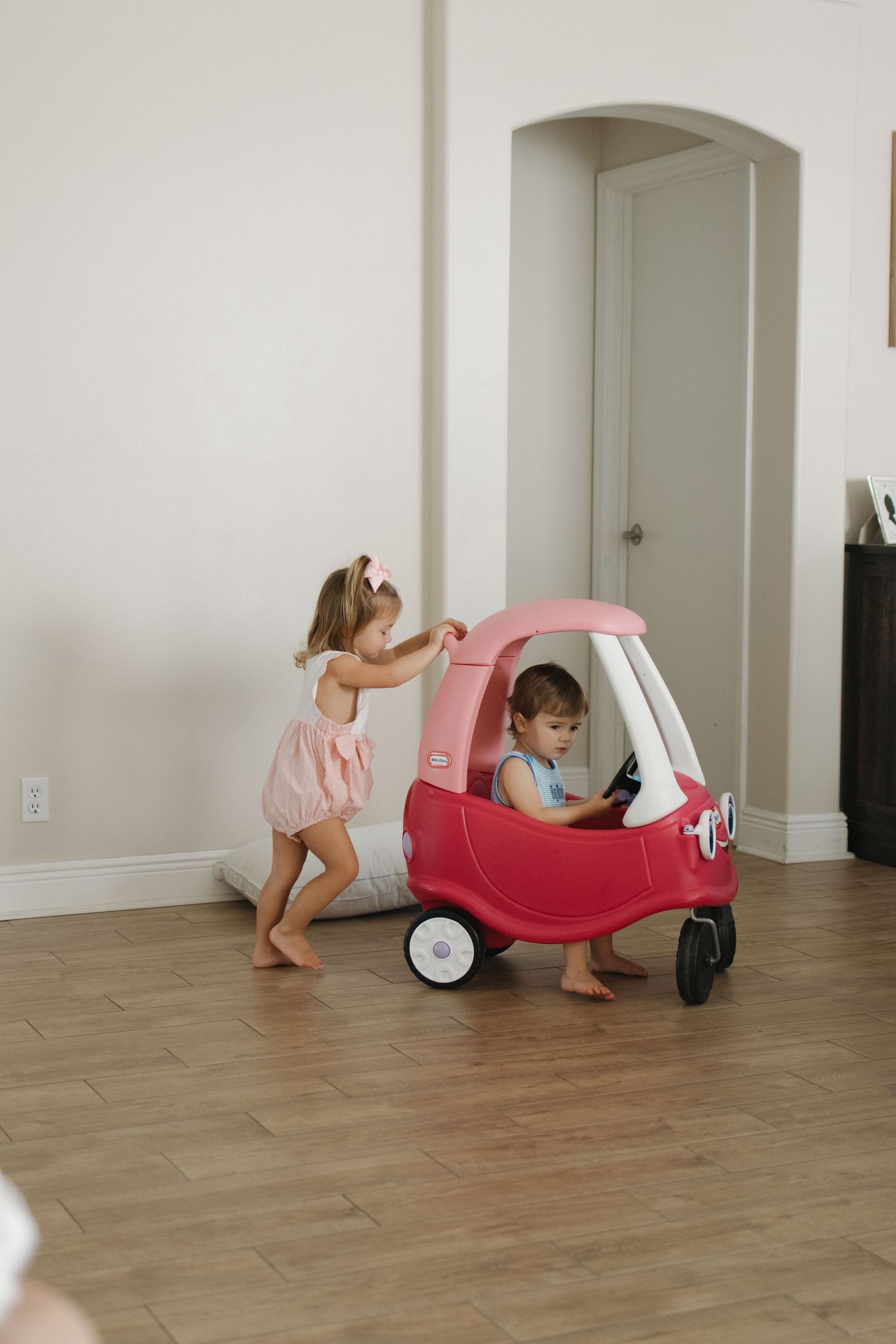 Children playing with a pink toy car on hardwood flooring.