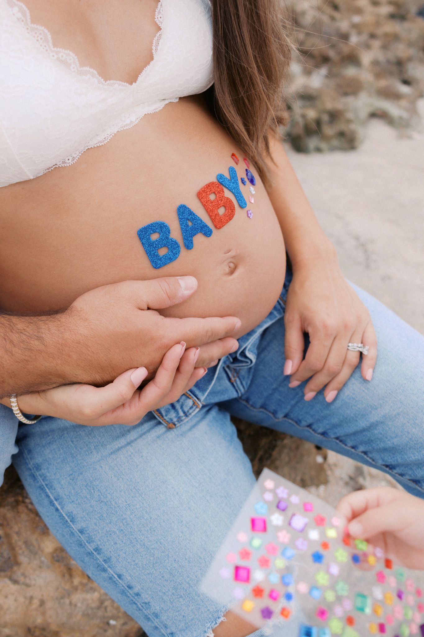 Pregnant woman with colorful BABY letter stickers on her baby bump, wearing white bra and jeans outdoors.