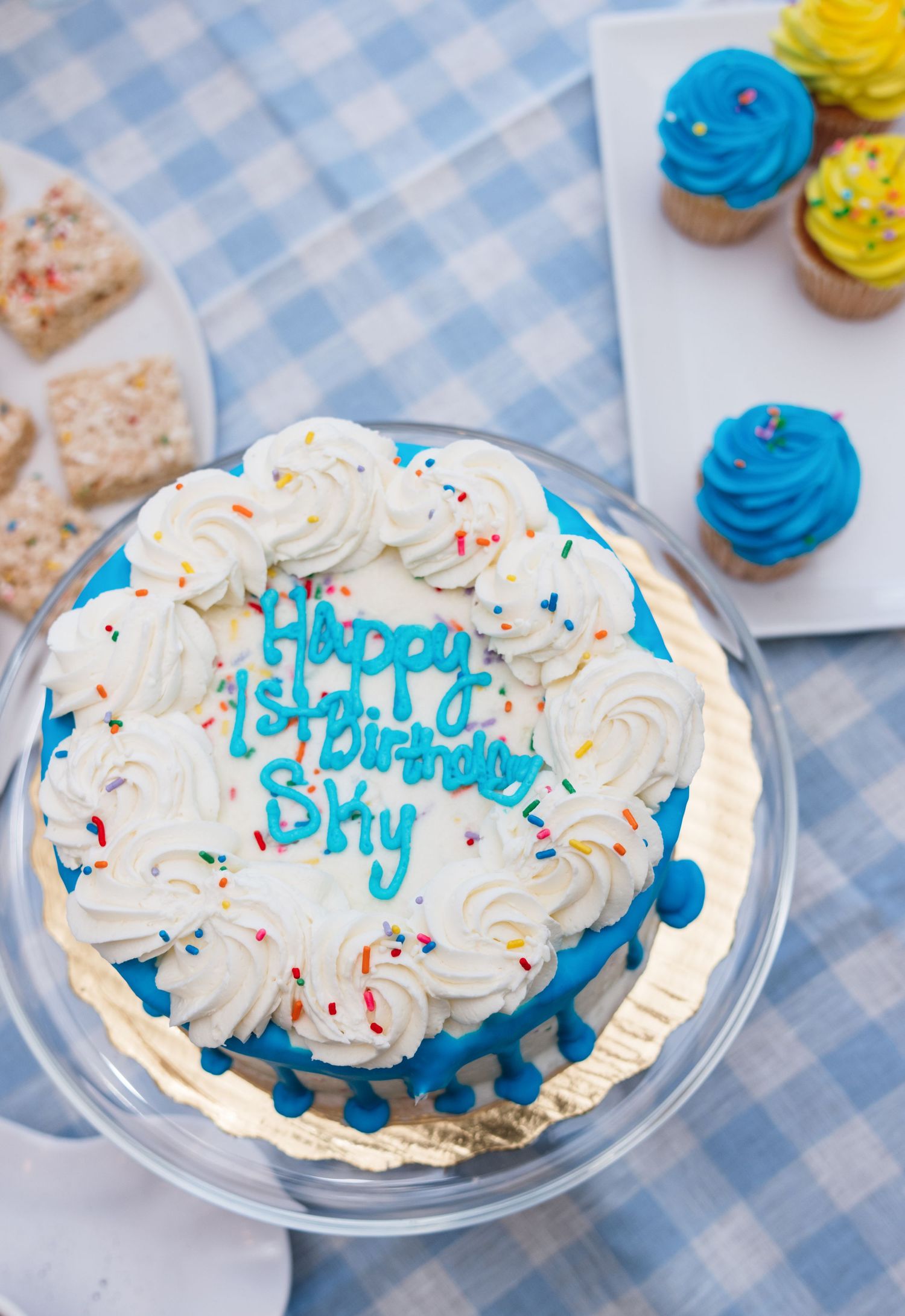 Birthday cake with blue and white frosting and sprinkles displayed on checkered tablecloth.