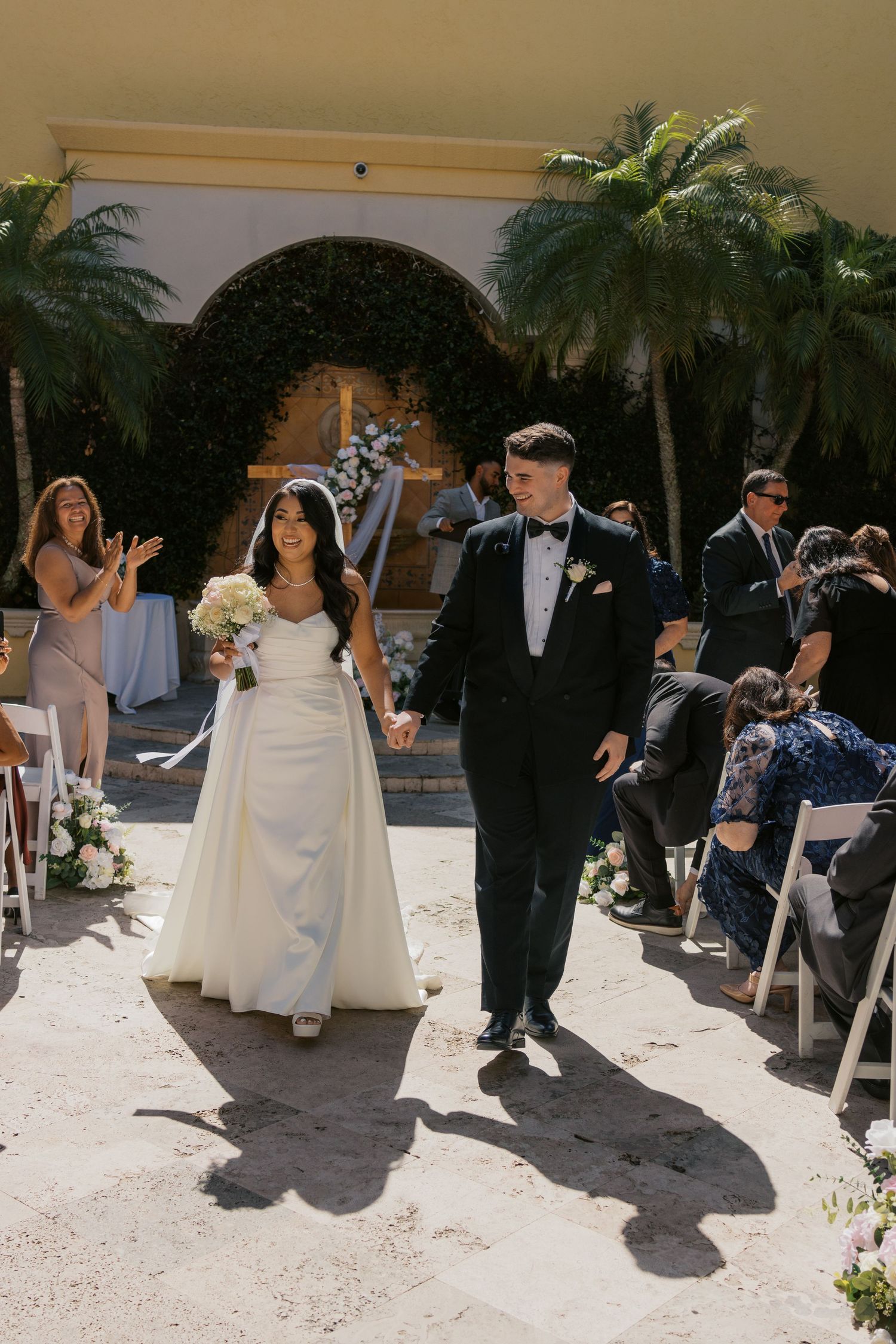 Bride and groom walk down the aisle after outdoor wedding ceremony, surrounded by guests and tropical palm trees.
