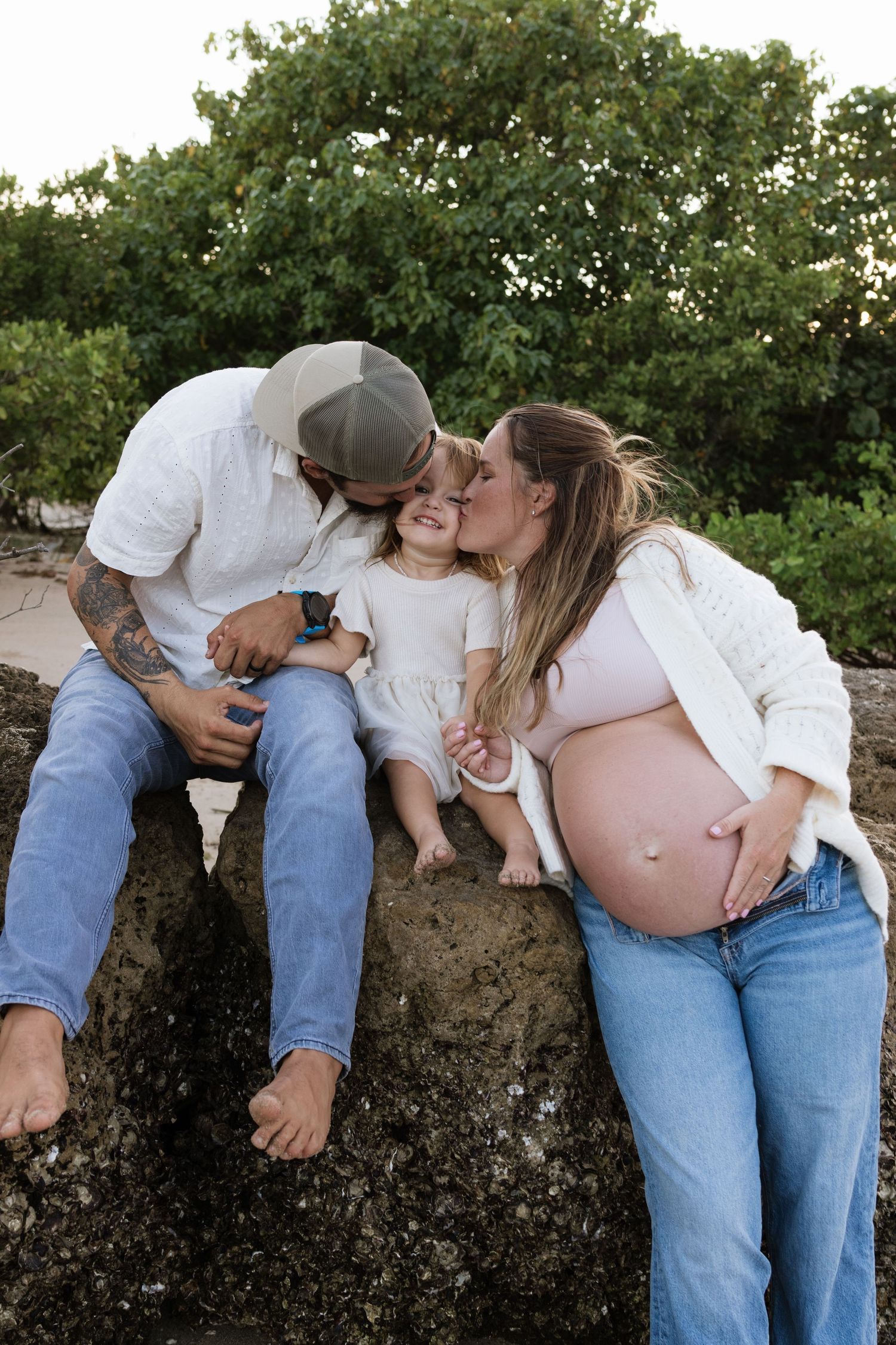Pregnant woman and tattooed man kiss their toddler daughter on cheeks while sitting on rocks outdoors.