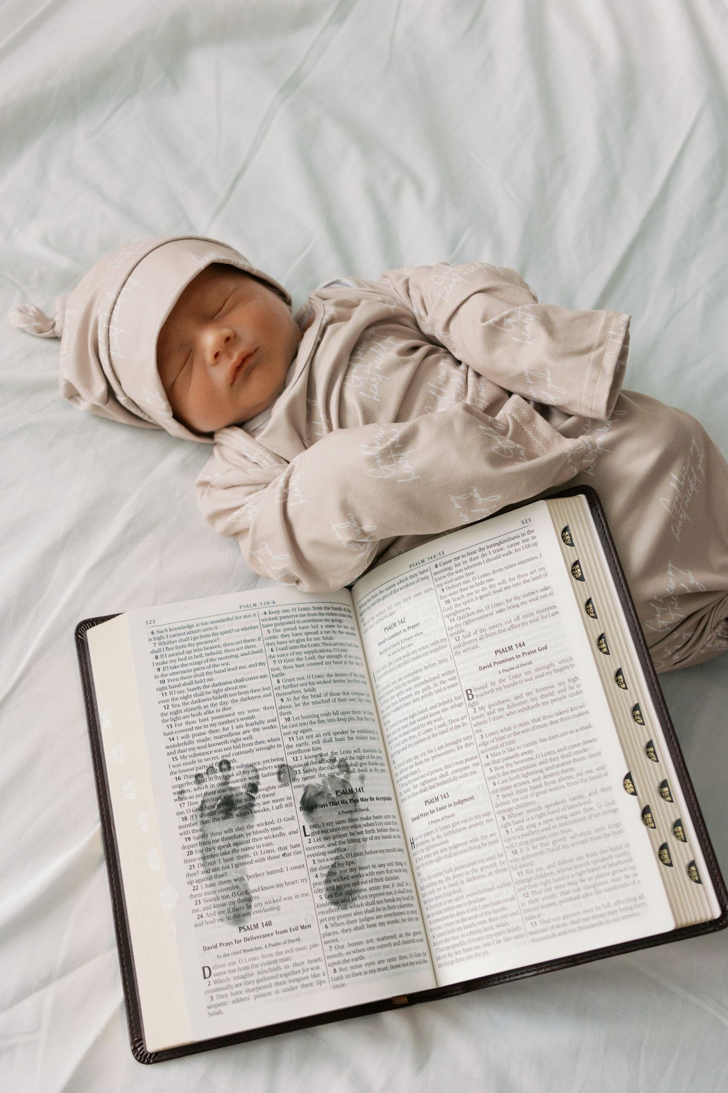 Newborn baby sleeping in beige outfit beside an open Bible with tiny footprints on the pages.